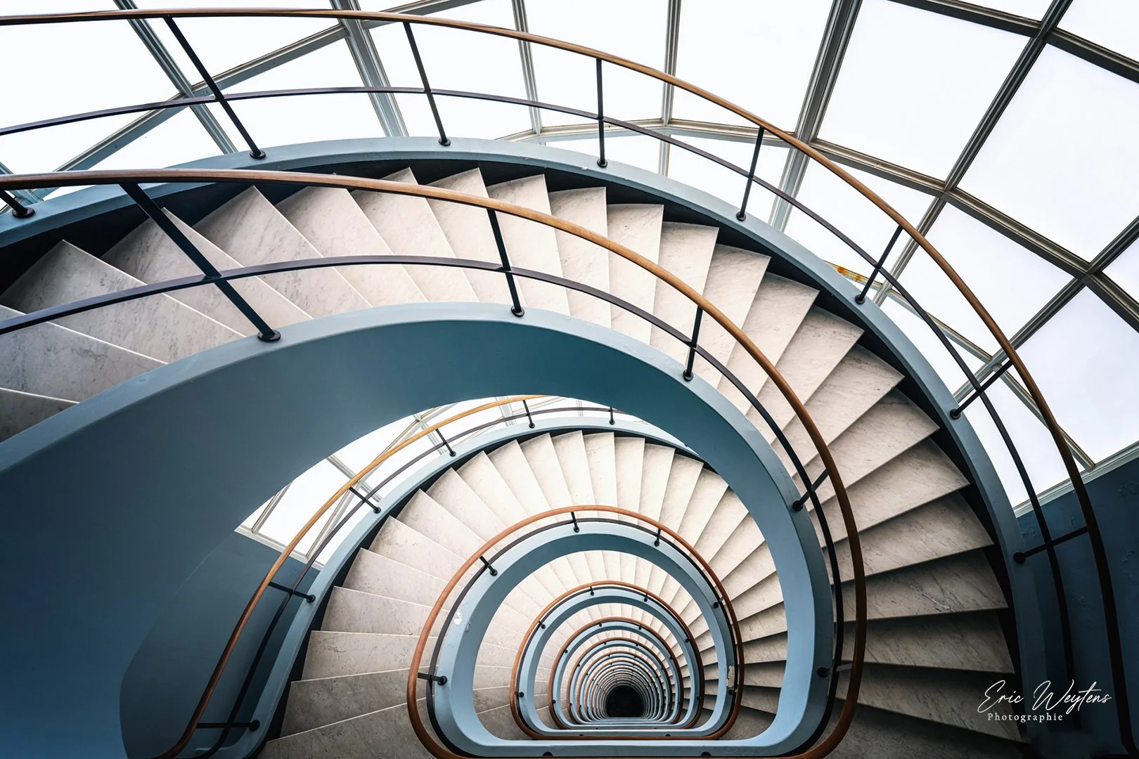 Rampe d'escalier en spirale vue du haut, avec des marches en pierre et des mains courantes en bois, entourée d'une structure en verre.