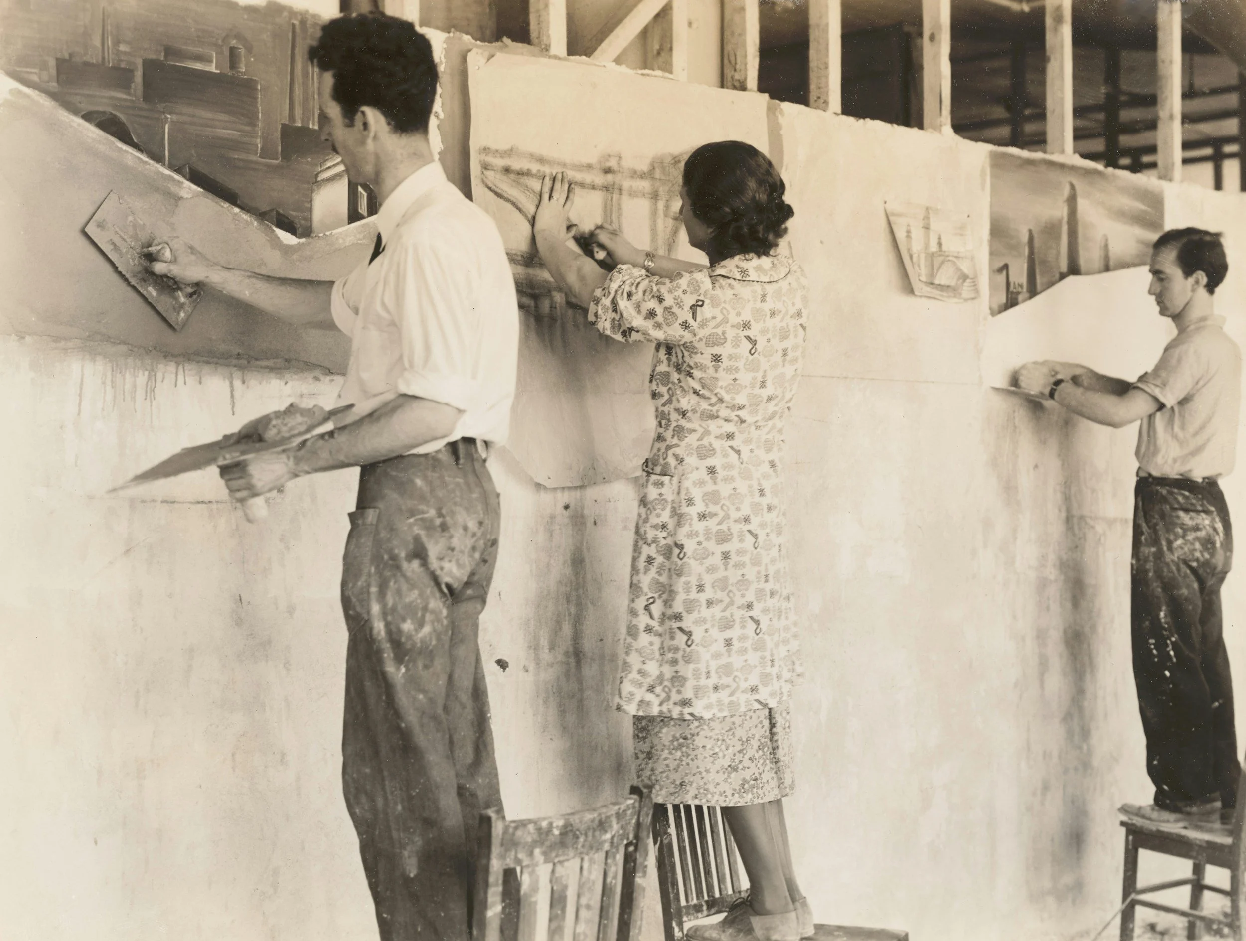 Three people painting a large mural or wall art, each standing on a chair, with the central woman painting a detailed landscape scene and the two men painting sections nearby.