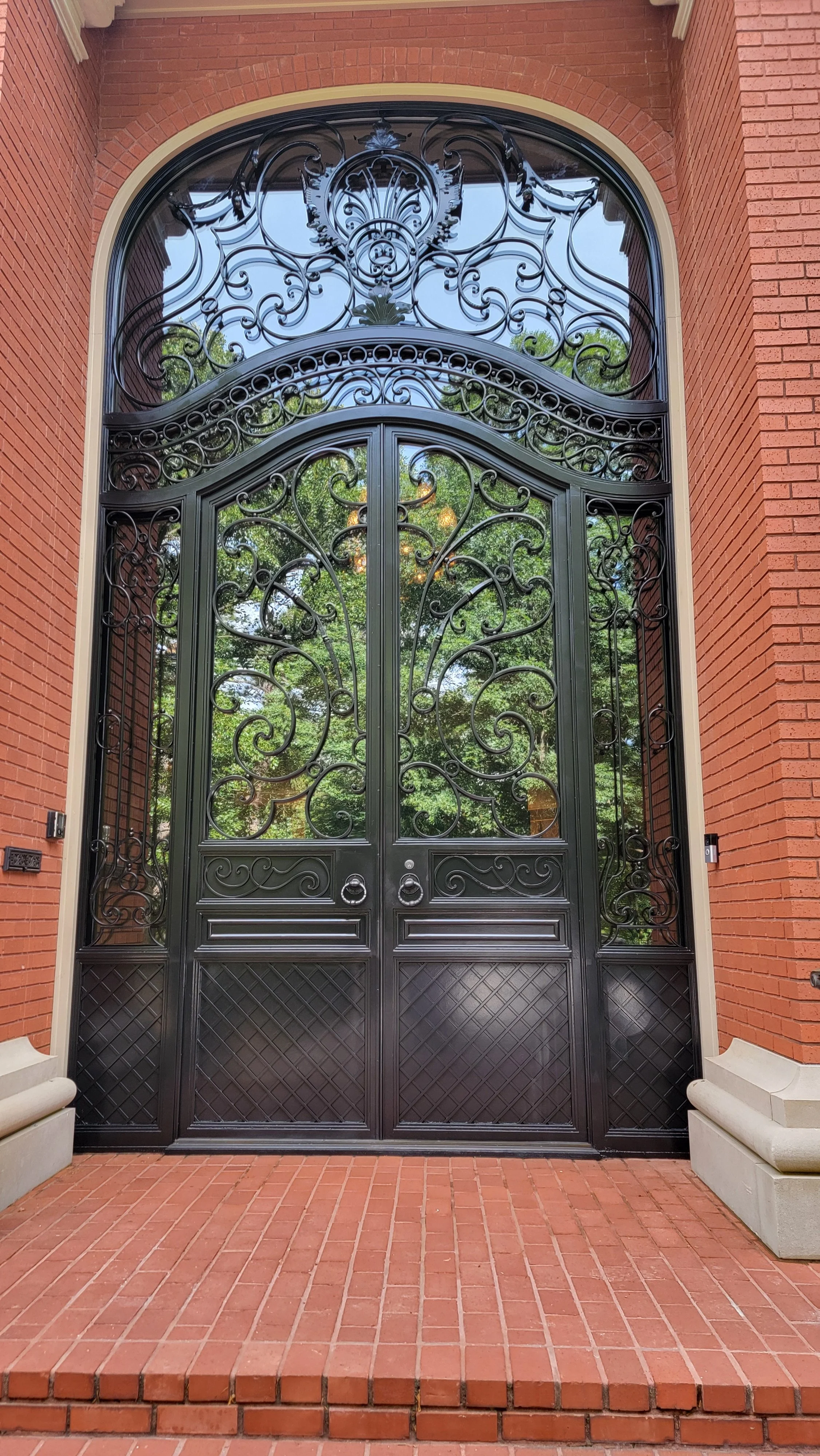 Ornate black iron gate with decorative scrollwork and glass panels, set within a red brick building entrance with stone steps and brick flooring.