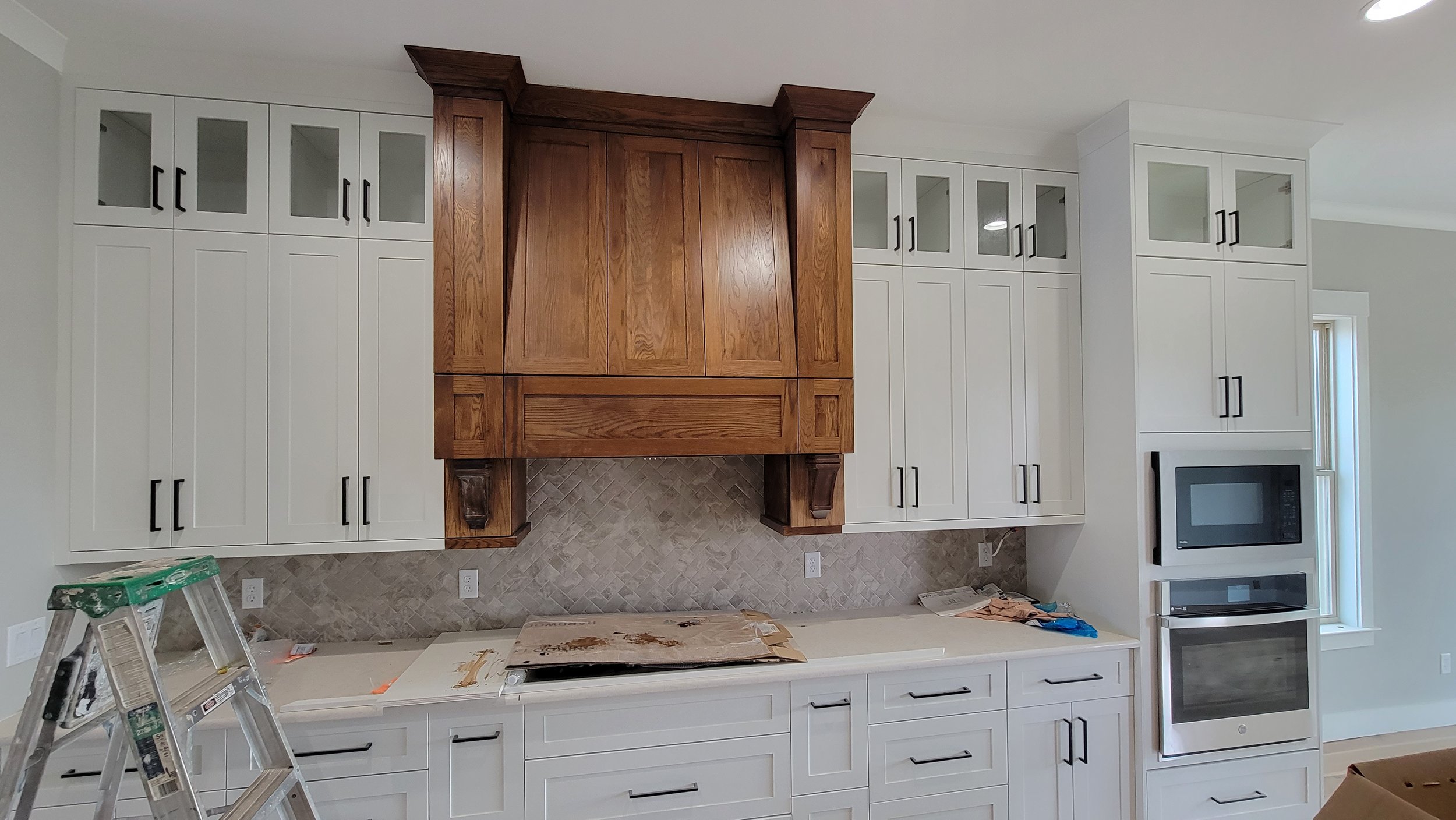 Kitchen under construction with white cabinets, a wooden range hood, tile backsplash, microwave, and oven.