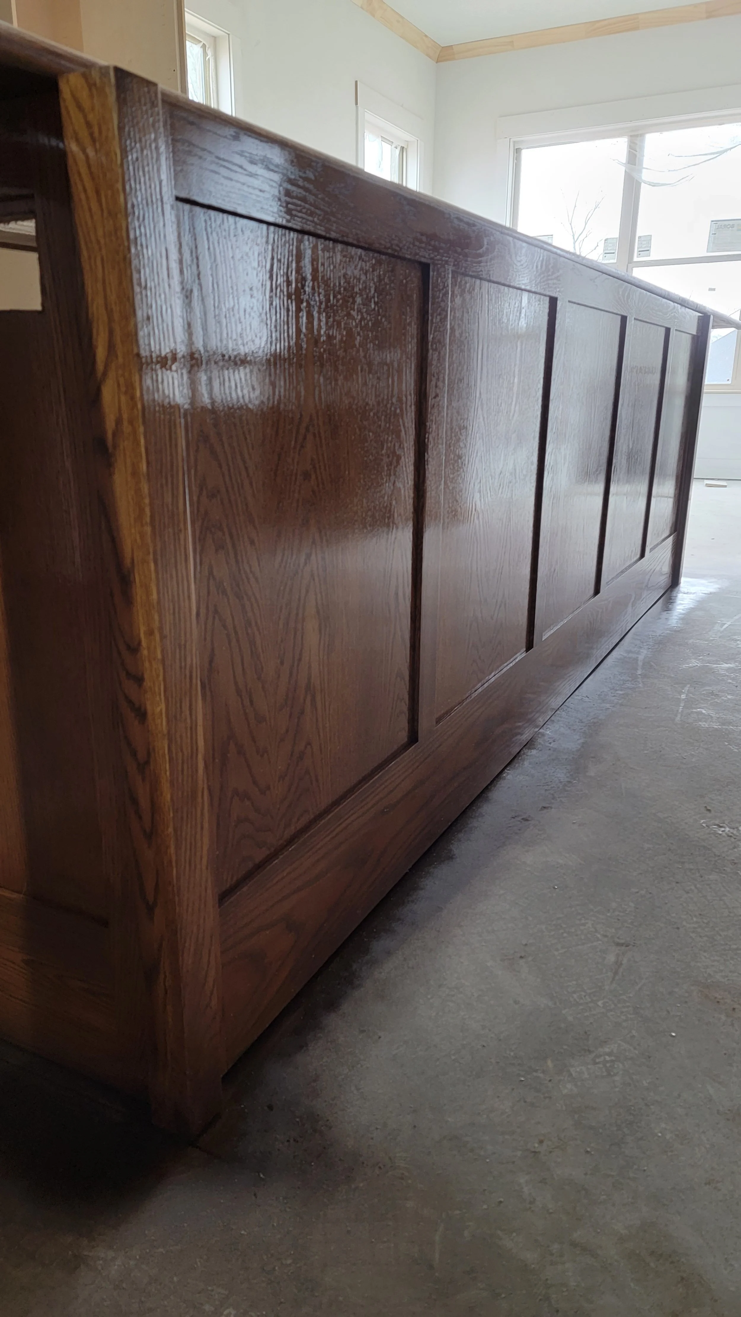 A close-up view of a polished wooden cabinet or sideboard in a room with large windows and a concrete floor.