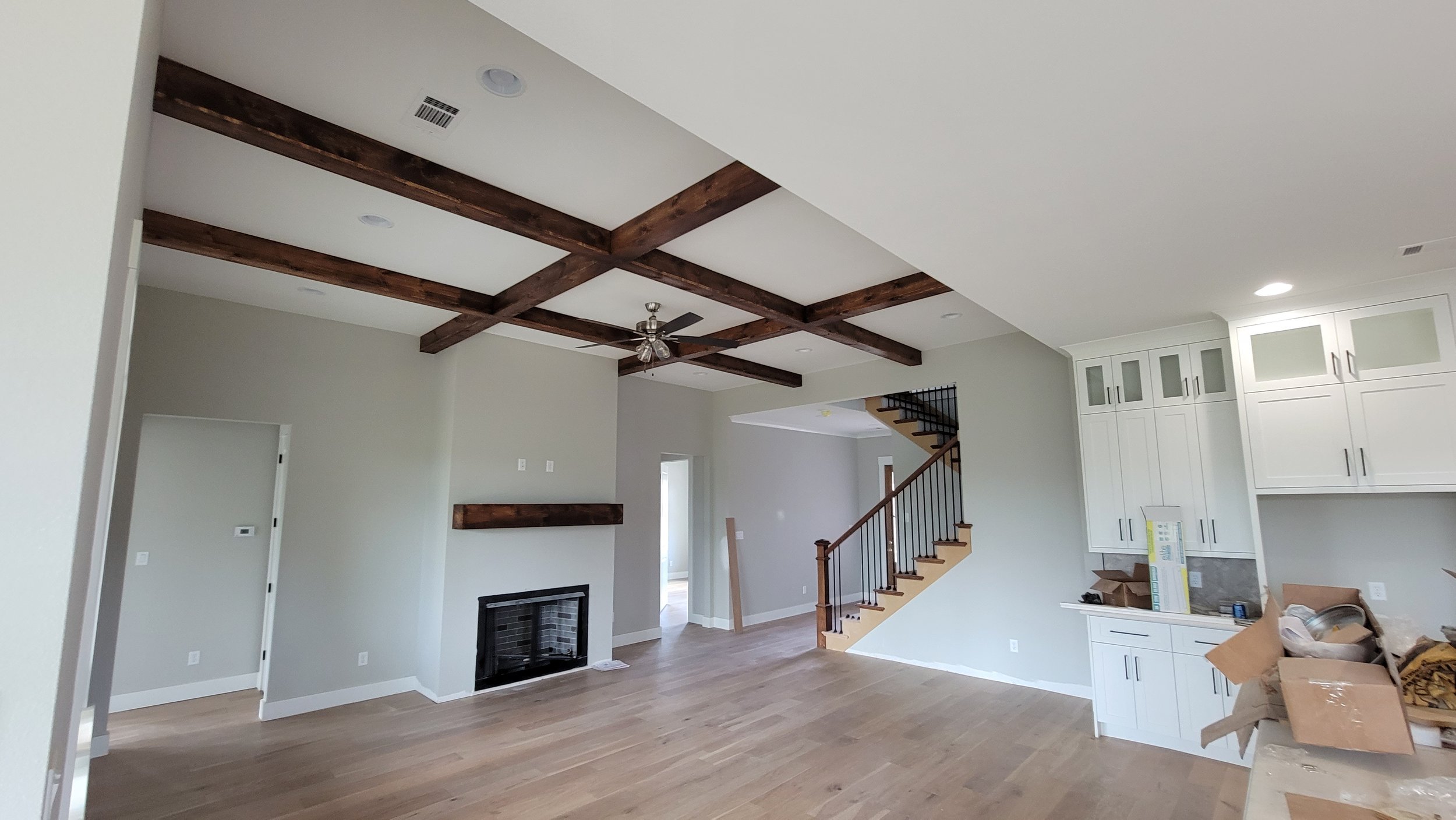 Empty living room with wood flooring, white cabinets in the kitchen area, a fireplace with a wooden mantel, and a staircase with wooden handrails and black balusters. The ceiling has dark wooden beams and a ceiling fan.