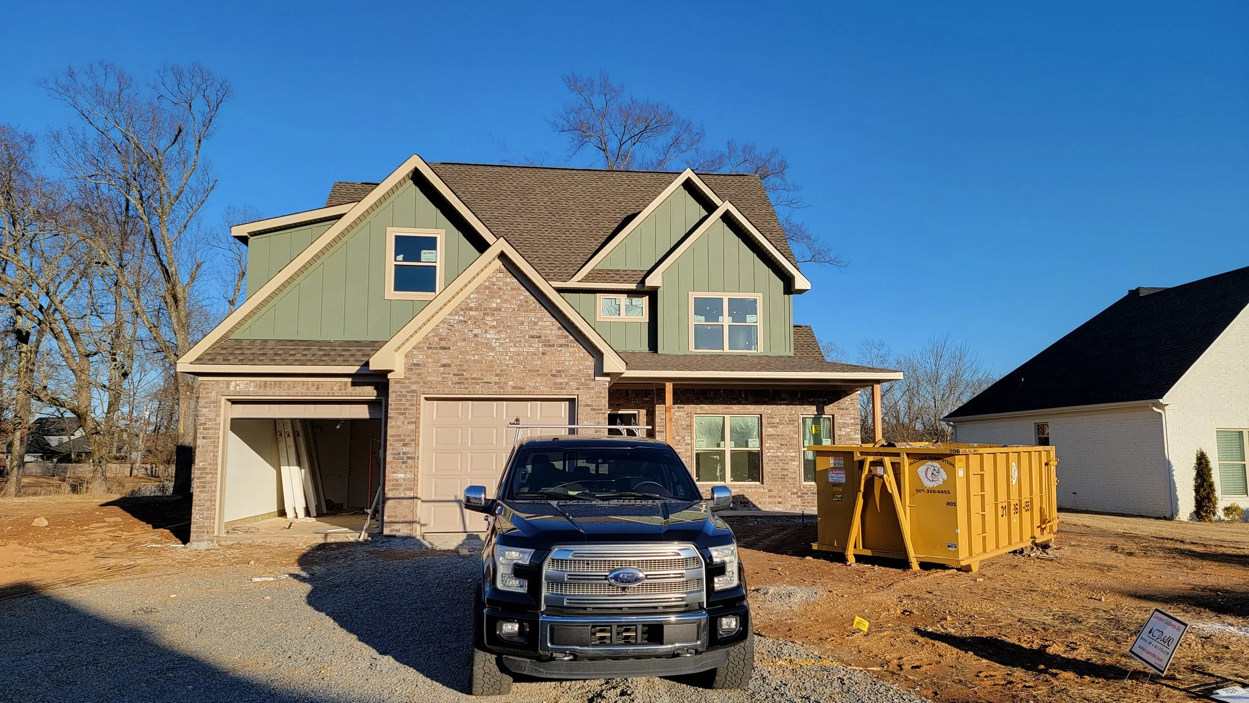 Newly built two-story house under construction with a black Ford truck parked in front and a yellow dumpster on the right side, clear blue sky, and neighboring house on the right.
