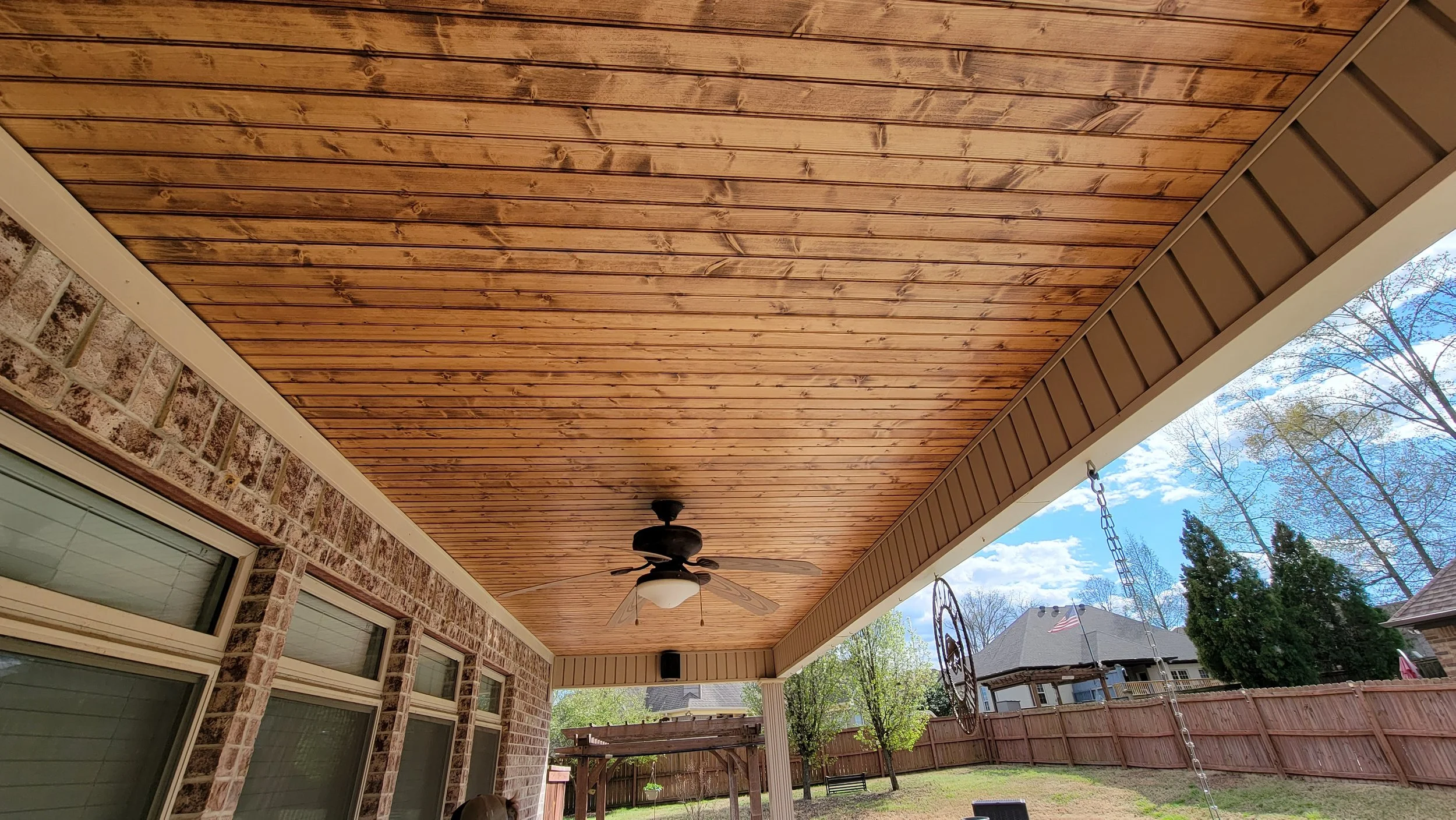 Covered backyard patio with a wooden ceiling, ceiling fan, brick exterior wall with large windows, wooden fence, swing set, and a blue sky with clouds.