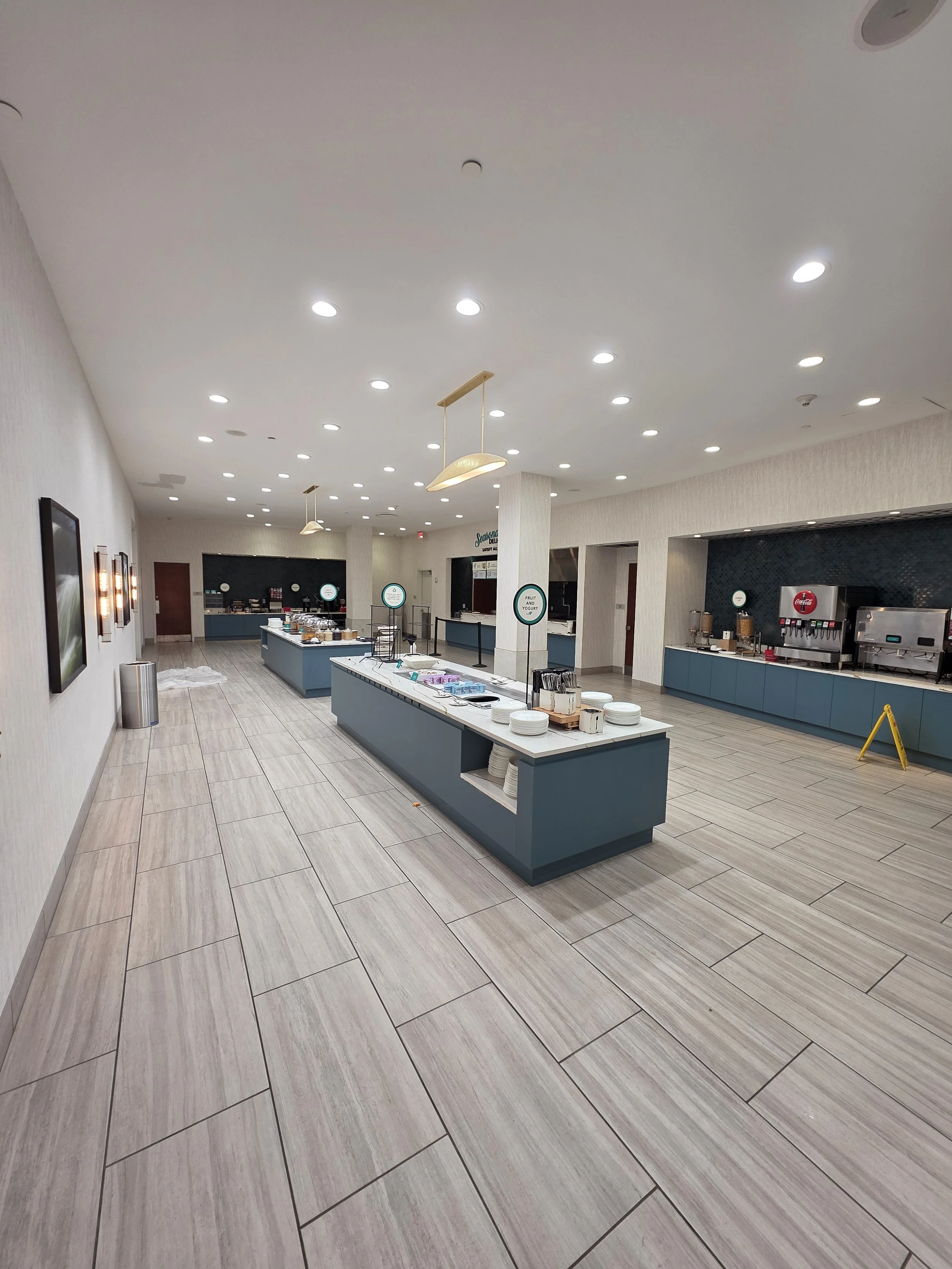 Empty breakfast buffet area with long counters holding plates, bowls, and utensils, surrounded by beige tiled flooring, white walls, and ceiling with multiple recessed lights and hanging lights.