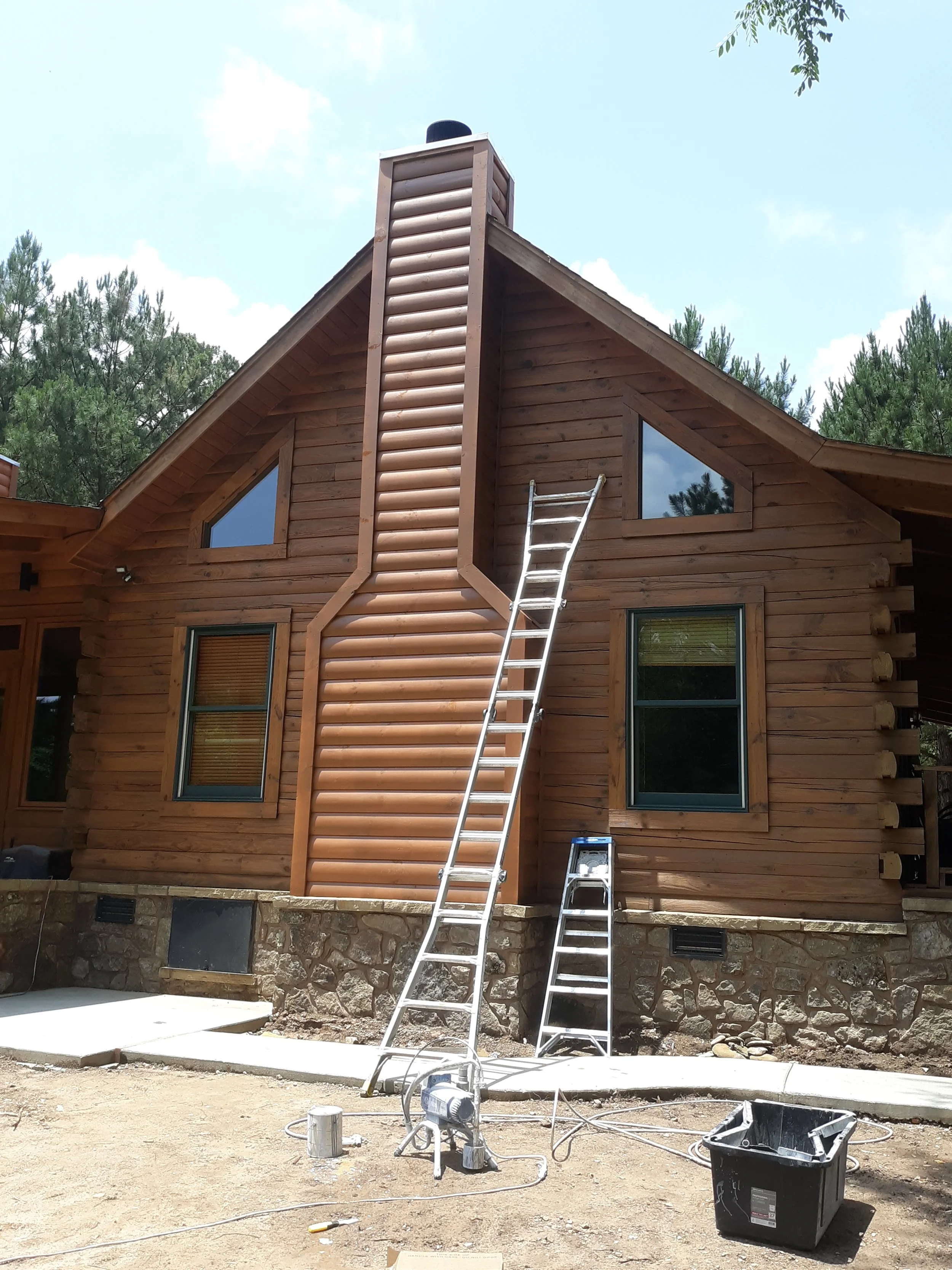 The image shows a wooden house under construction with ladders leaning against it, a chimney, and construction tools on the ground, with trees and a partly cloudy sky in the background.