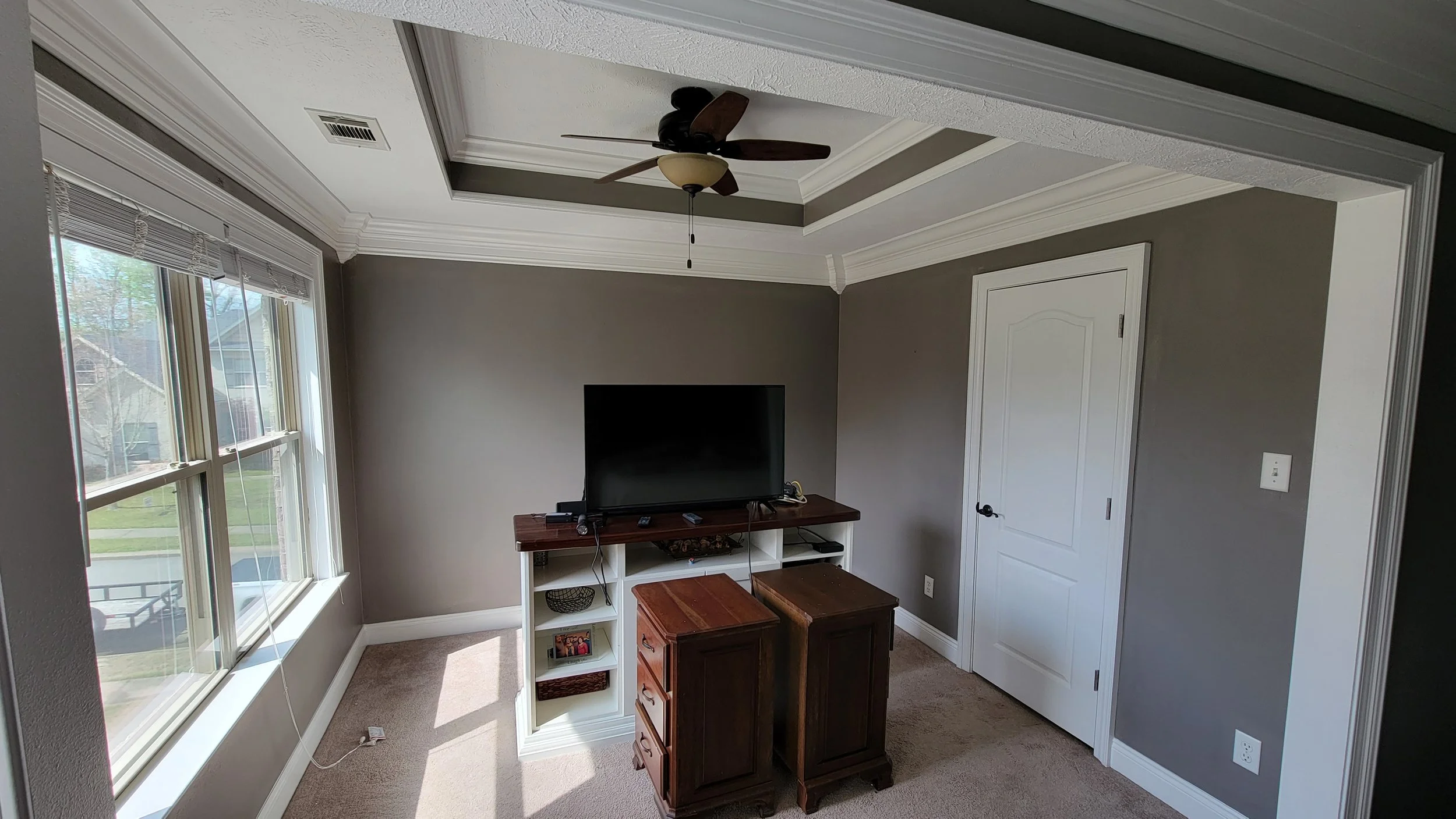 Living room with large window, gray walls, white crown molding, ceiling fan, a flat-screen TV on a white wooden stand, and two small brown wooden cabinets in front of the TV.