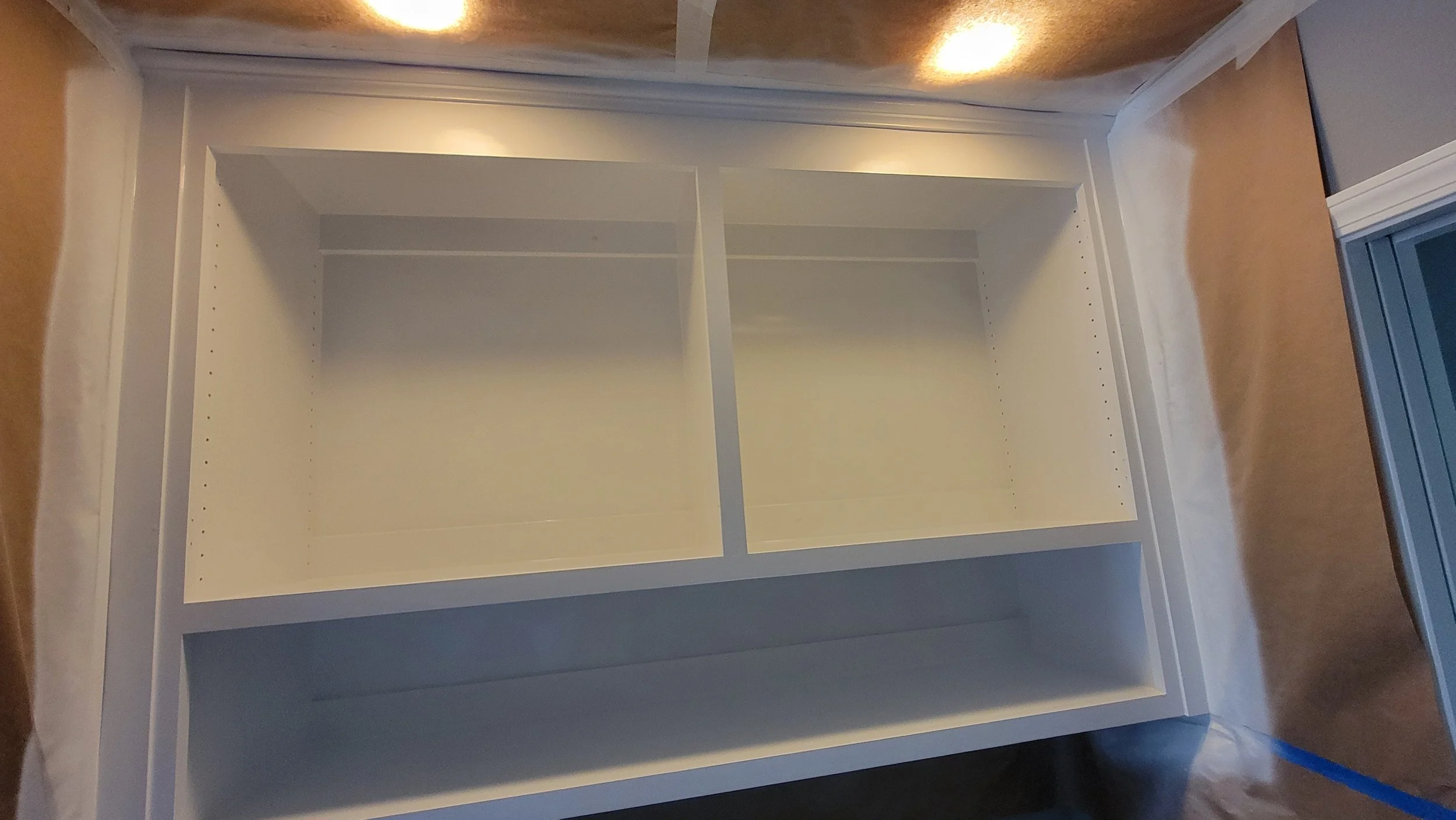 White built-in shelving unit with three shelves, partially unassembled, in a room with tan flooring and blue painter's tape on the floor.