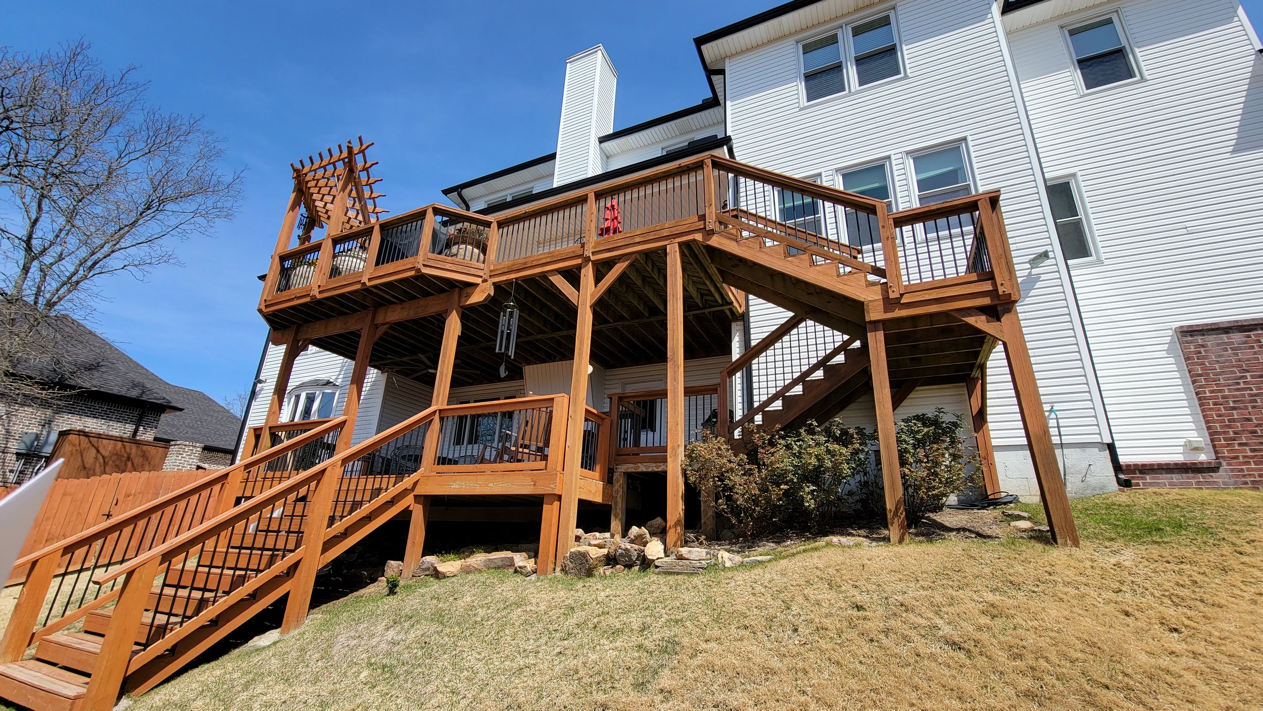 Wooden deck with stairs attached to a white house, featuring a railing and a lattice pergola on top,, with a grassy yard and neighboring houses in the background.