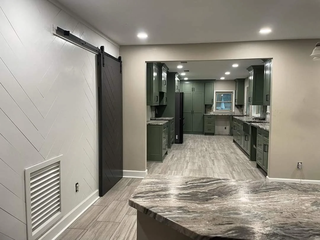 Modern kitchen with green cabinets, granite countertops, and gray wood-look flooring, viewed through an open archway.