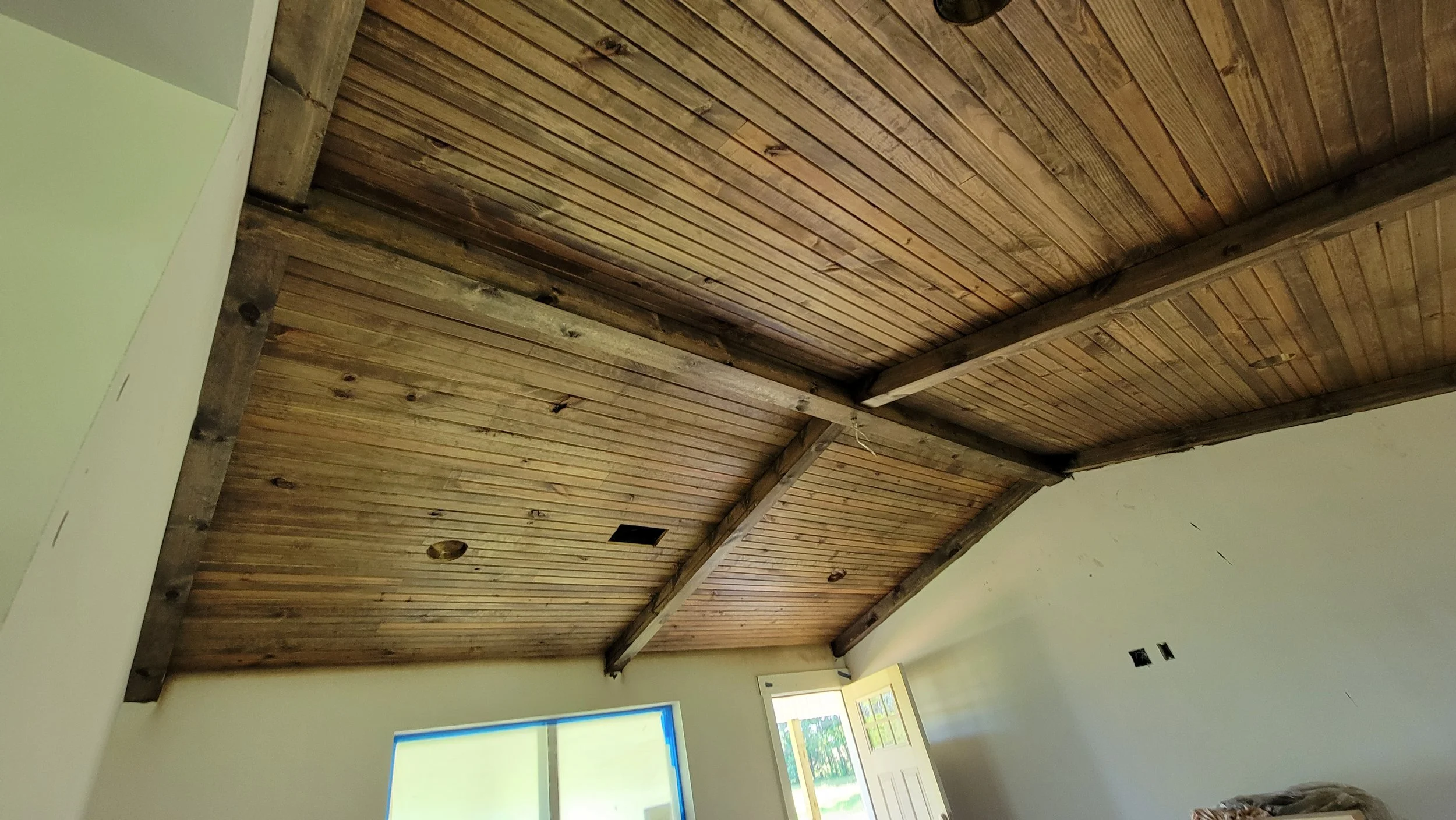Interior of a room with a wooden ceiling, partially finished walls, a window with blue tape around it, and an open door leading outside, with a view of greenery.