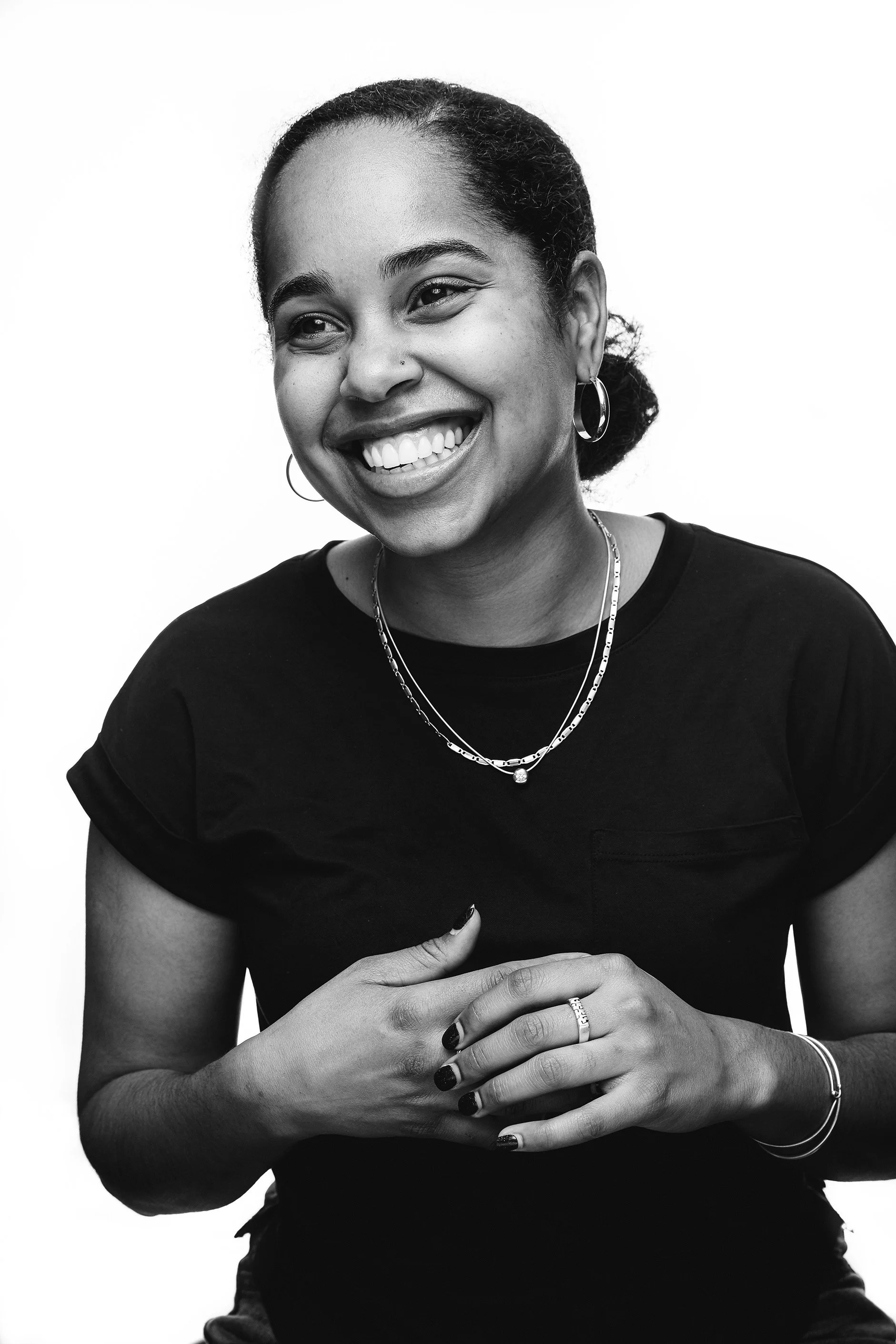 Black and white portrait of a young woman smiling, wearing hoop earrings, layered necklaces, and a black t-shirt, with her hands clasped together.
