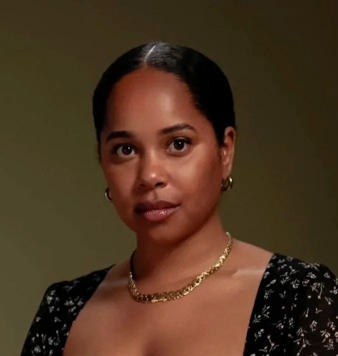 A woman with dark hair pulled back, wearing gold hoop earrings, a gold necklace, and a black floral top, looking directly at the camera with a neutral expression against a plain background.