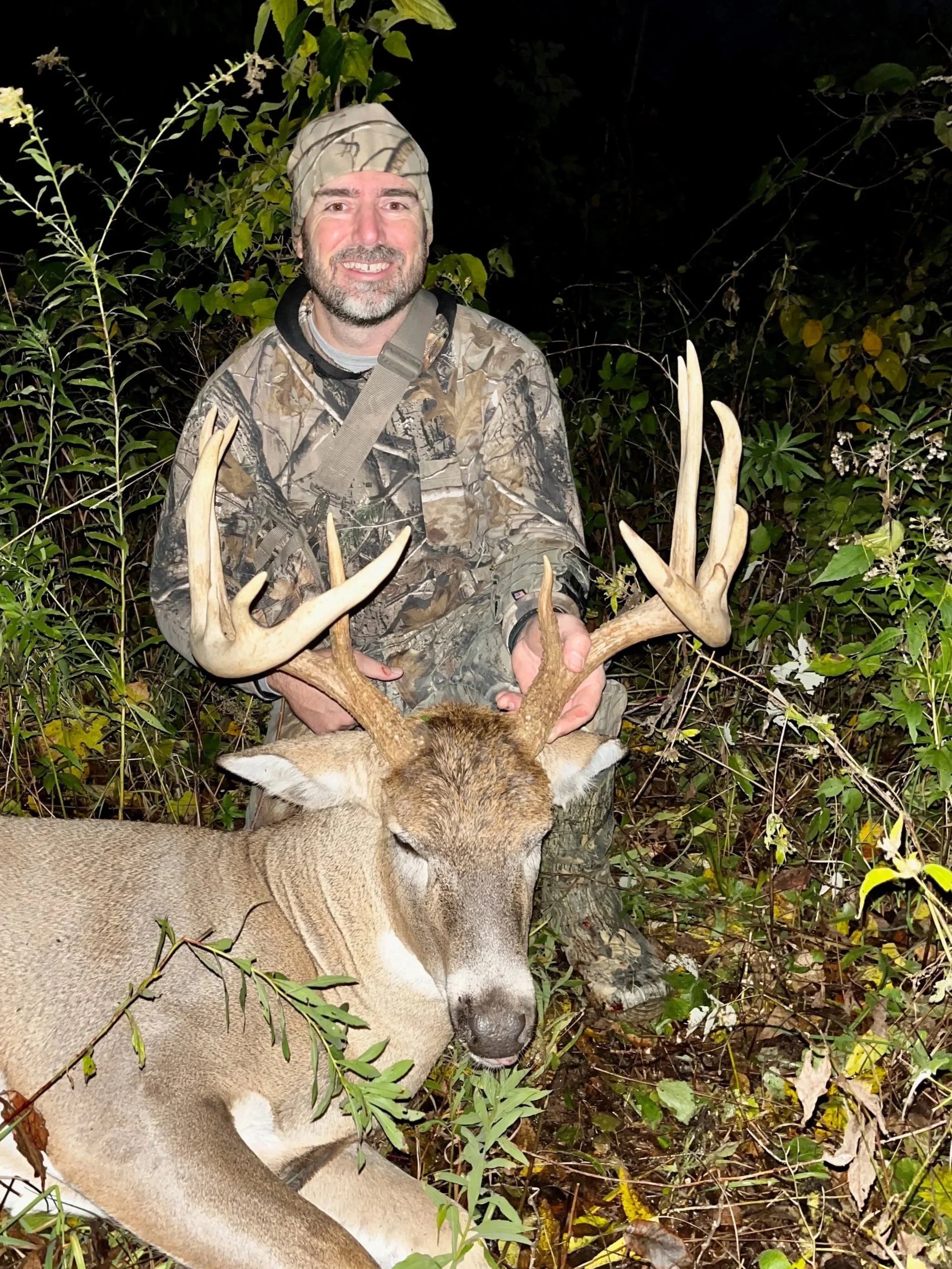 A man in camouflage clothing holding a large deer with antlers in a forest at night.