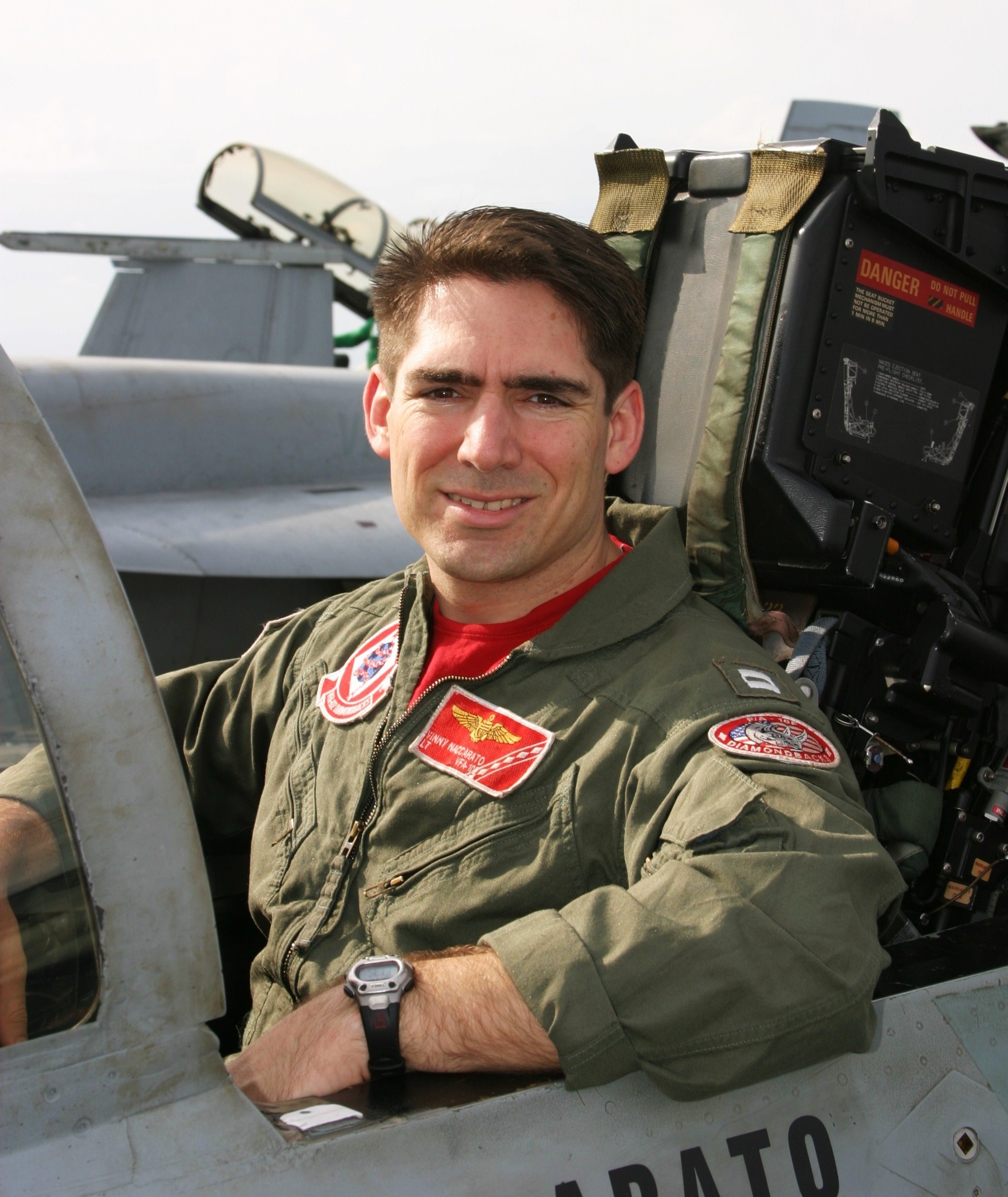 A male pilot with short, dark hair in a military fighter jet cockpit, wearing a green flight suit with patches, a red T-shirt underneath, and a black watch.
