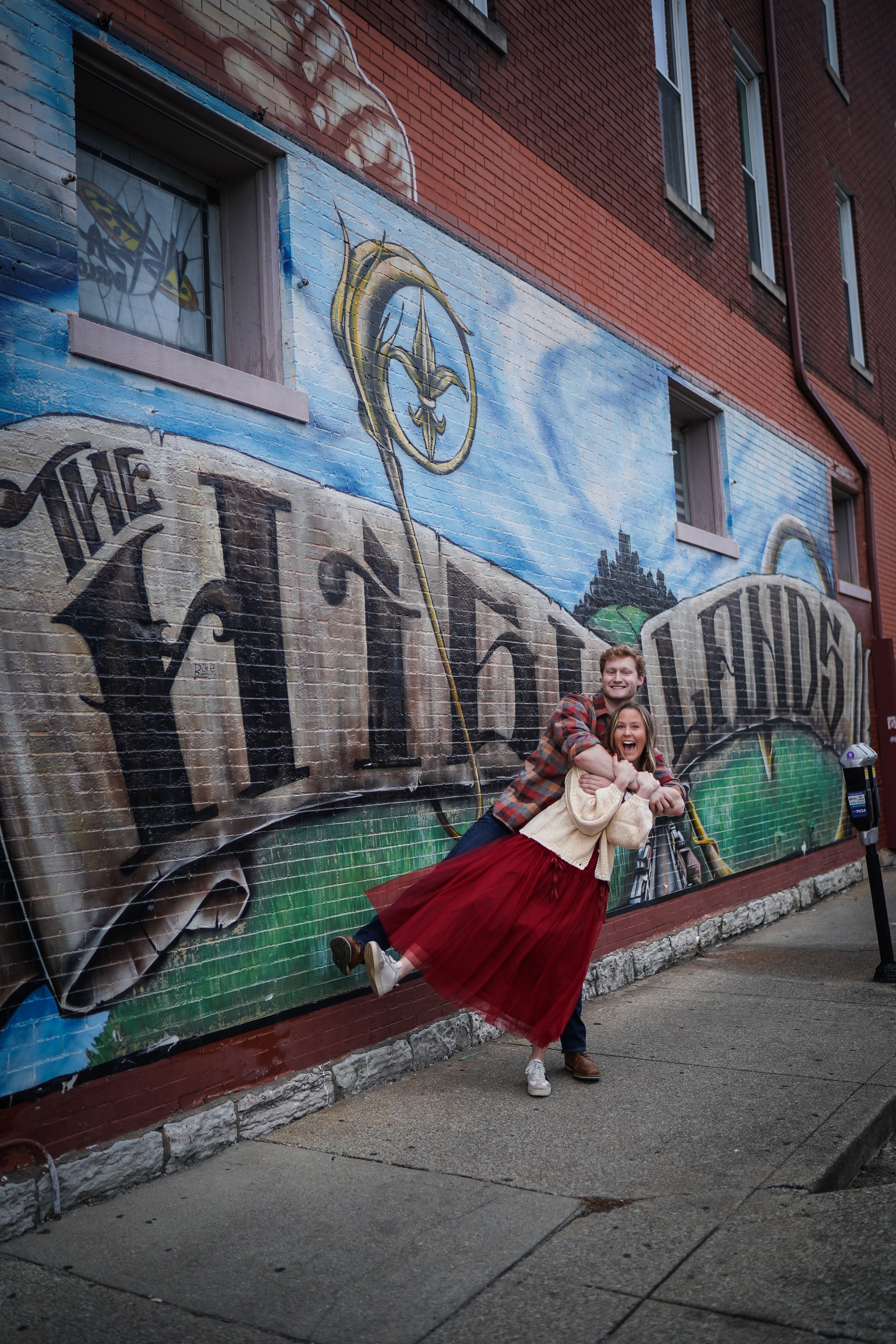 Two people, a man and a woman, playfully hugging in front of a colorful mural on a brick wall, with the woman leaning back with her arms around the man's shoulders and the man holding her around the waist. The mural features large street art lettering and a design resembling a medieval key or scepter.