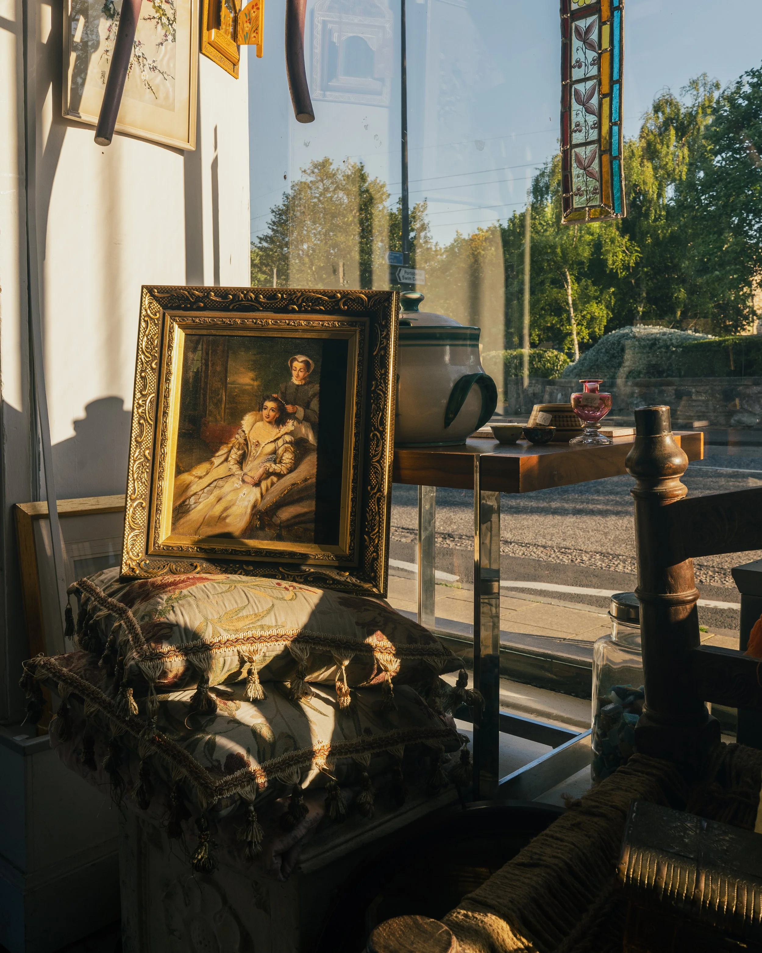 Vintage art and decorative items inside a store window, including a framed painting of a woman, textile cushions, a large teapot, jewelry, stained glass piece, and various small objects, with trees and a street visible outside.
