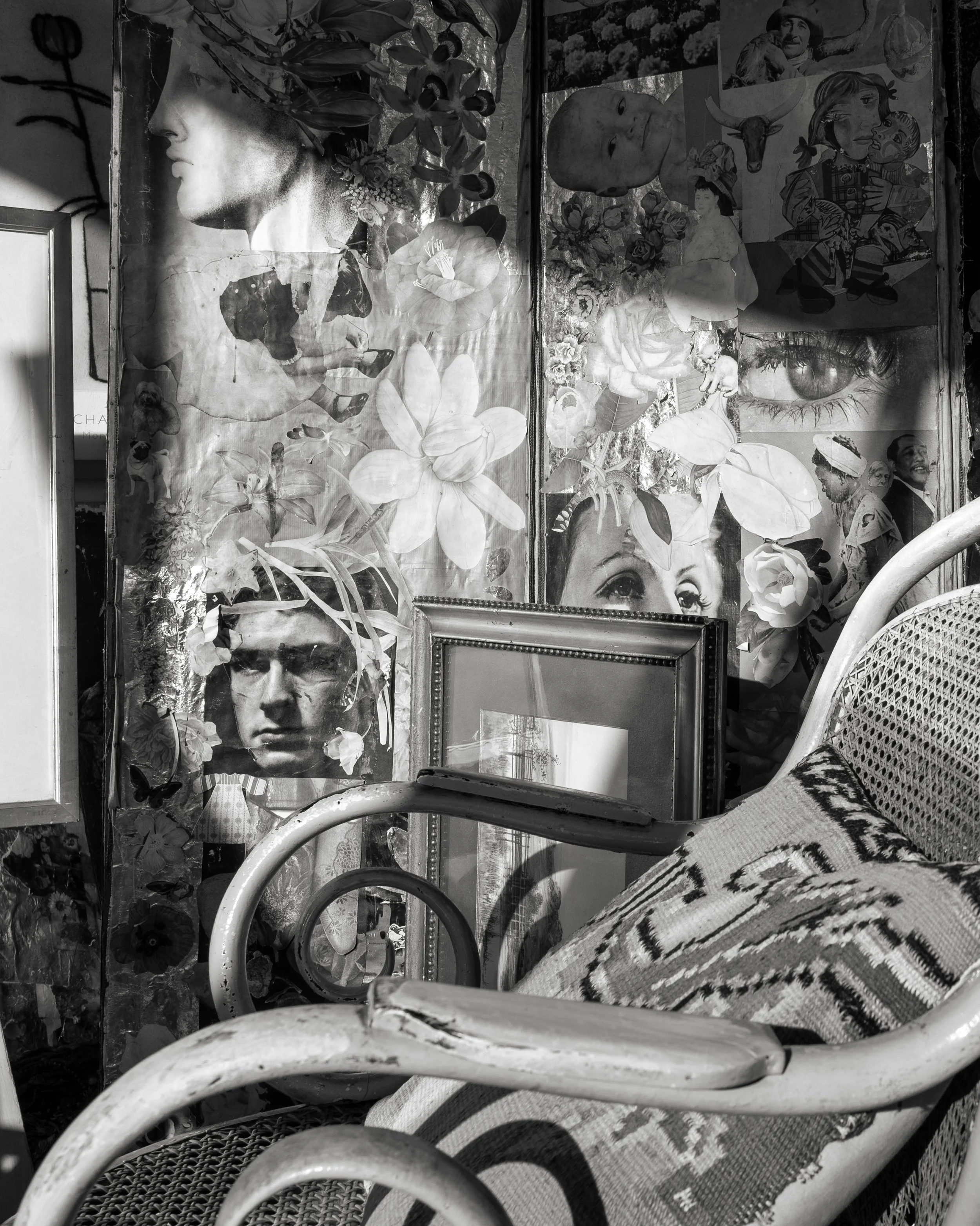 Black and white photo of a collage on a wall with various images including a woman's profile, a man's face, flowers, a large eye, a girl, and other pictures. There's a vintage chair with a patterned cushion and a framed mirror leaning against the collage.