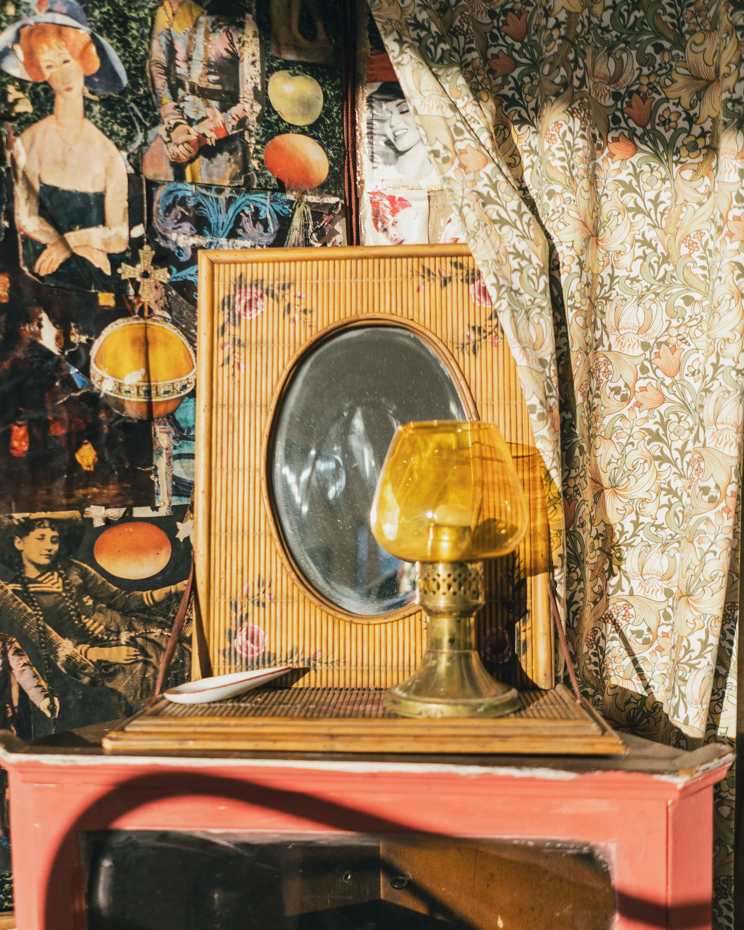 A vintage setting with a small round mirror framed with bamboo, a yellow amber glass lamp, a white dish, and a patterned curtain and wall covered with old photographs and magazine images, creating an eclectic, nostalgic atmosphere.