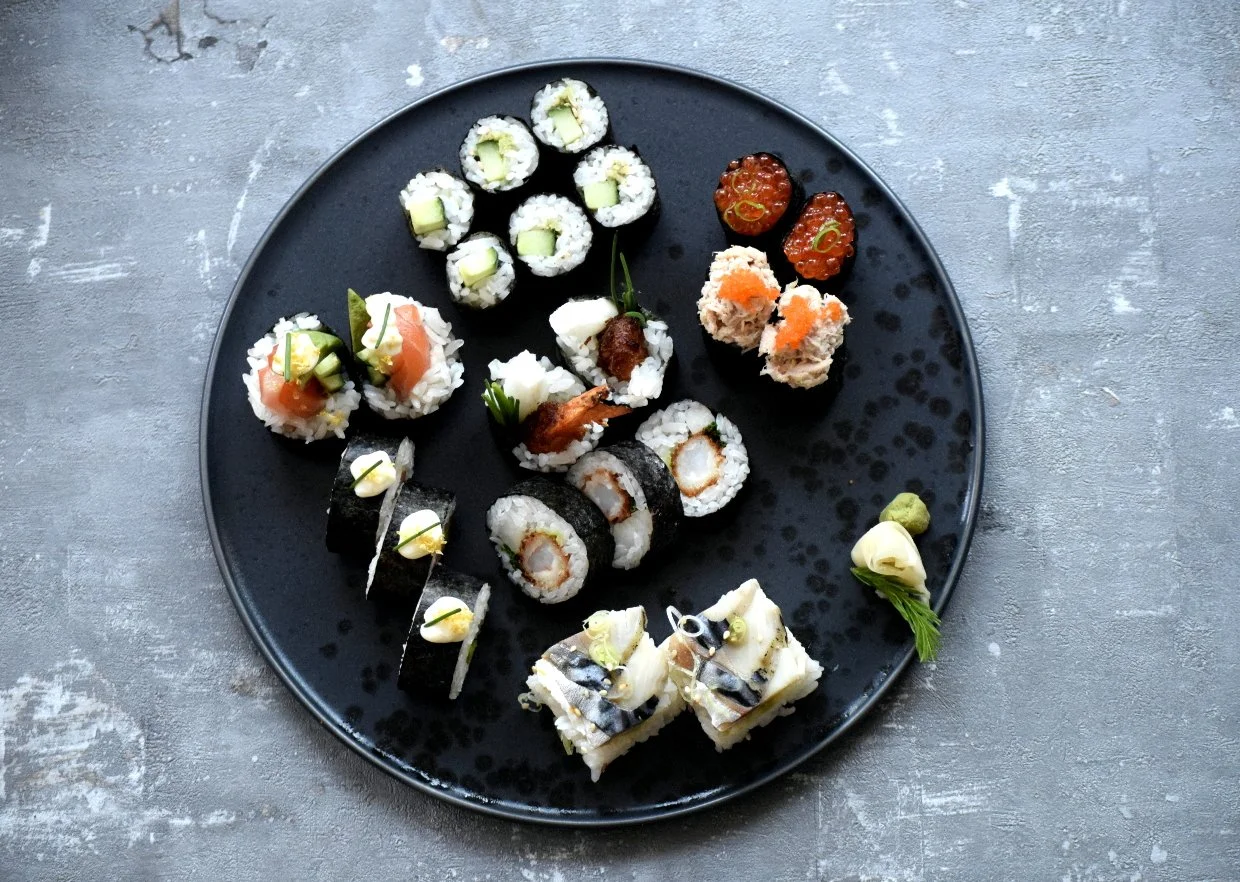 A black plate with a variety of sushi rolls, garnished with wasabi, ginger, and soy sauce, taken from Kimura's kitchen cookbook