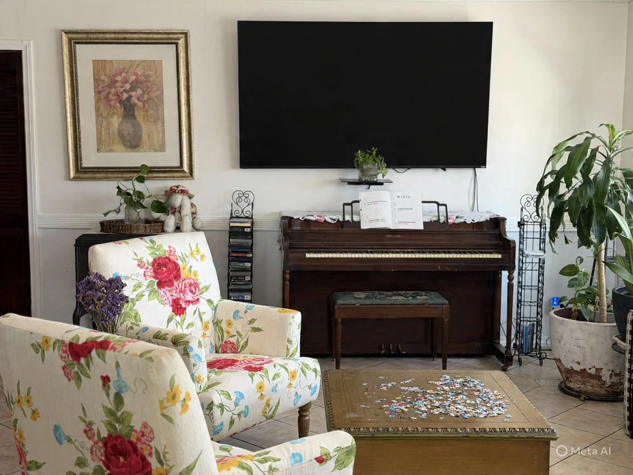 Living room with floral armchairs, a wooden table with puzzle pieces, and a piano against the wall. Potted plants and artwork decorate the space.