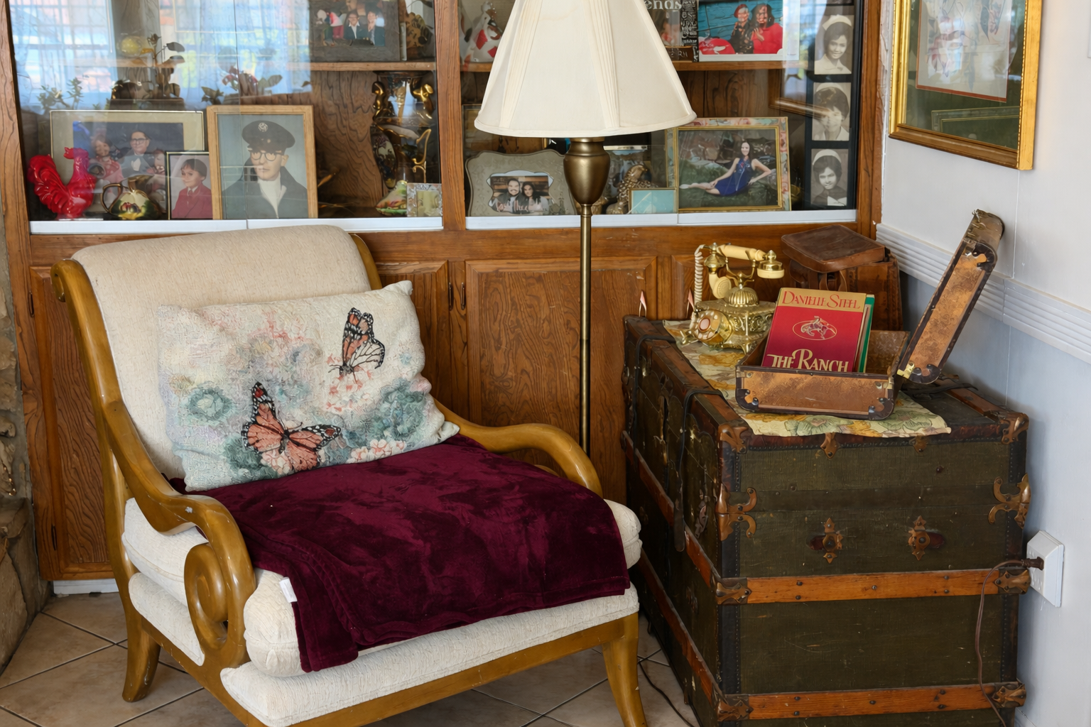 A vintage-style sitting area with a cream-colored armchair that has a butterfly-embroidered pillow and a burgundy throw blanket. Next to the armchair is a closed old trunk used as a table, holding a gold telephone, a small wooden box, and a vintage s