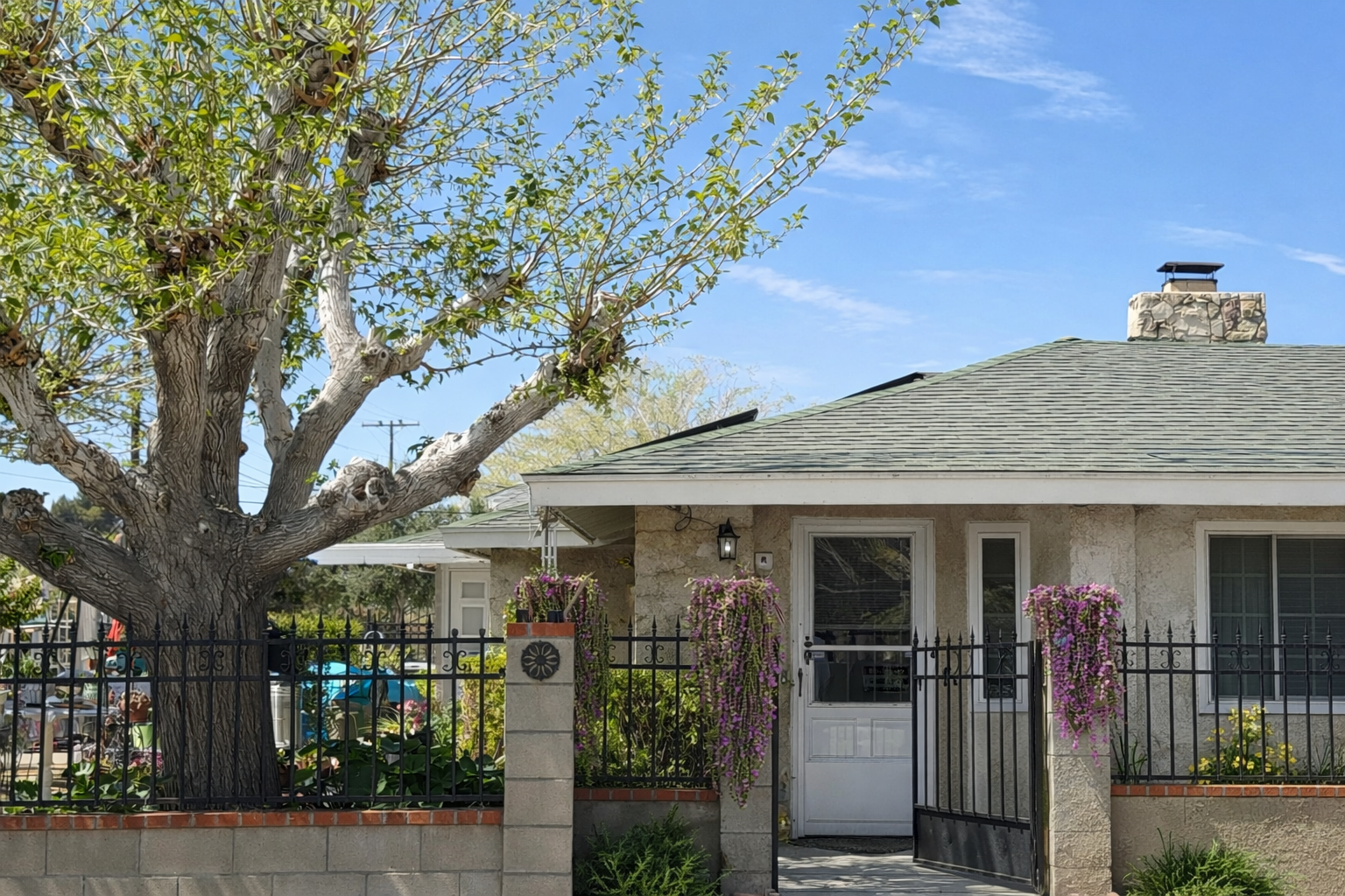 House with a tiled roof, stone exterior, front door, surrounded by a black metal fence, with hanging pink and purple flowers on the posts, a large tree in front, and a clear blue sky.