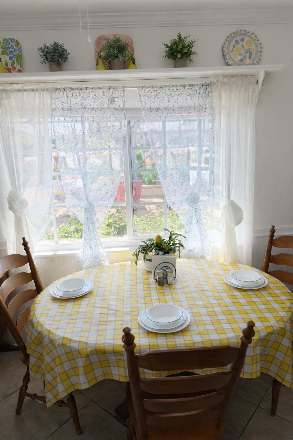 A cozy dining area with a round table covered in a yellow and white checkered tablecloth, four white plates, a potted plant, and a window with sheer curtains overlooking a garden.