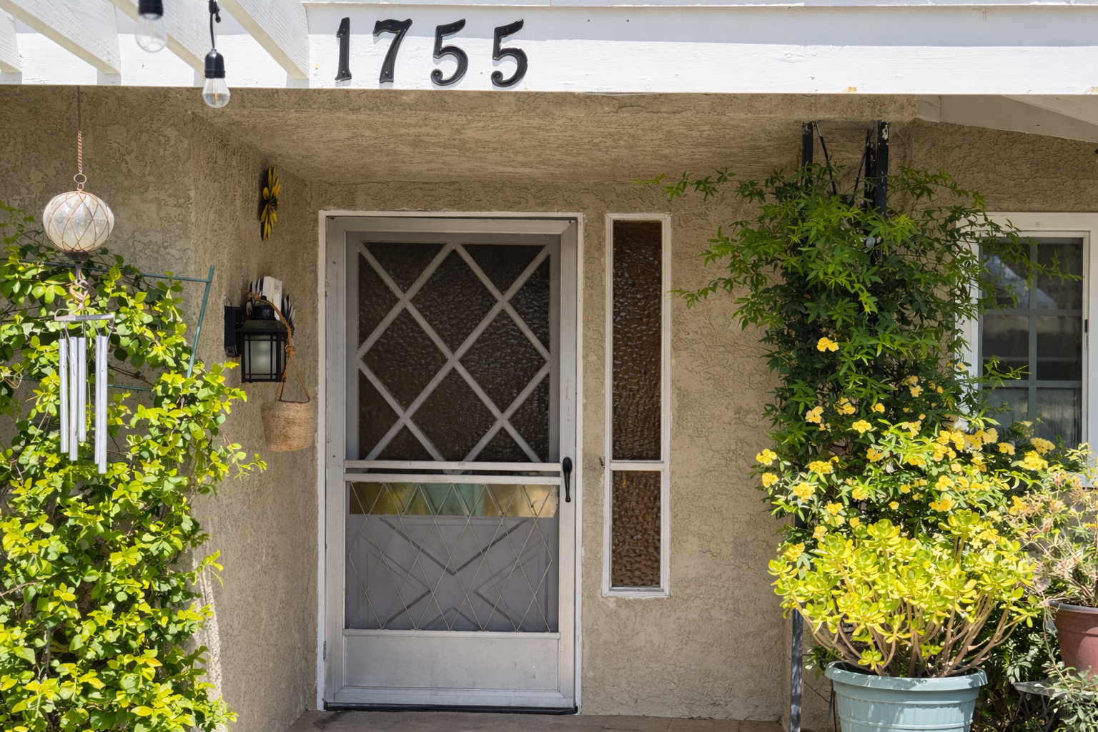 Front door of a house with beige stucco walls, flanked by potted plants with yellow flowers, a wind chime, and a lantern-style light fixture, with house number 1755 displayed above.