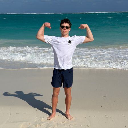 Young man standing barefoot on the beach, flexing biceps, wearing sunglasses, a white t-shirt, and navy shorts, with ocean and sky in the background.
