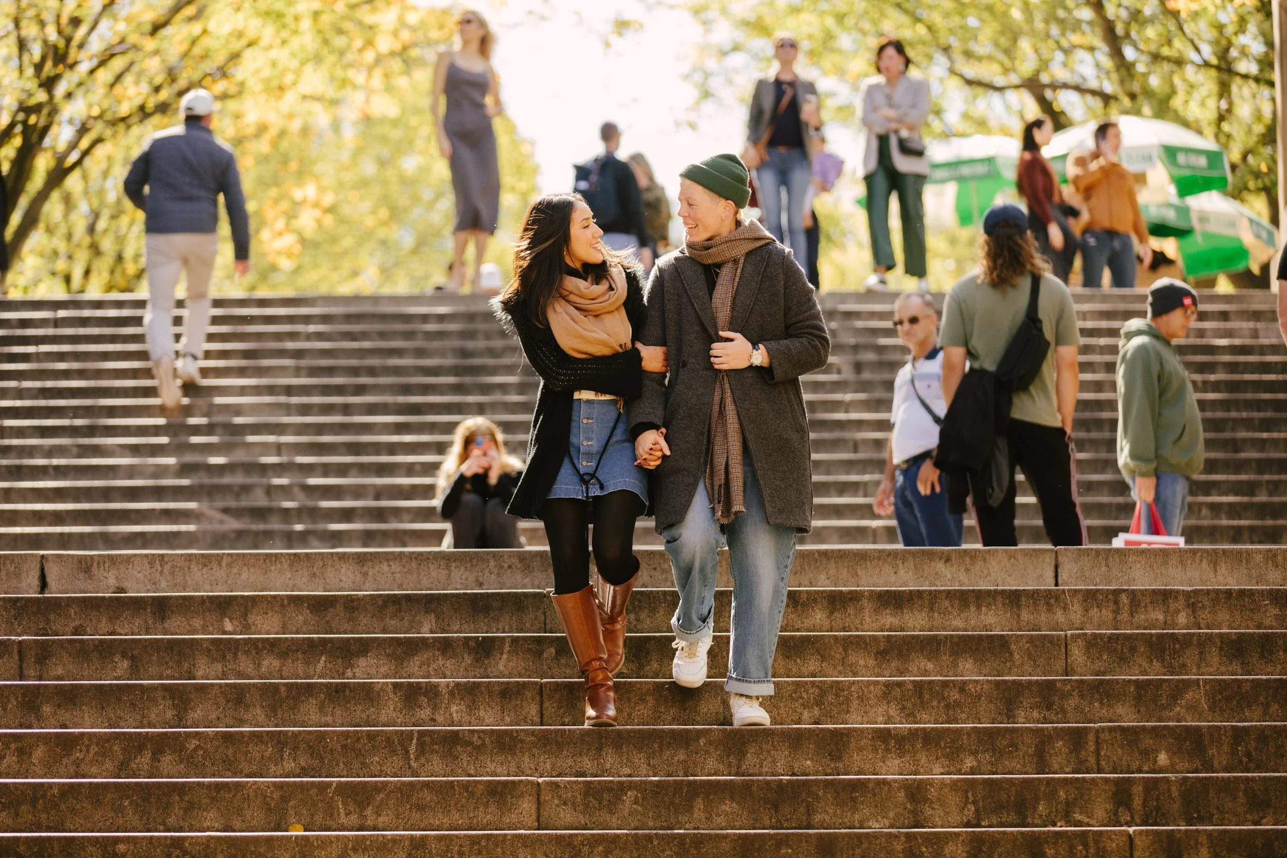 Two young women walk up outdoor stairs, holding hands, smiling, in autumn clothing, with multiple people on the stairs and at the top, in a park with trees and sunlight in the background.