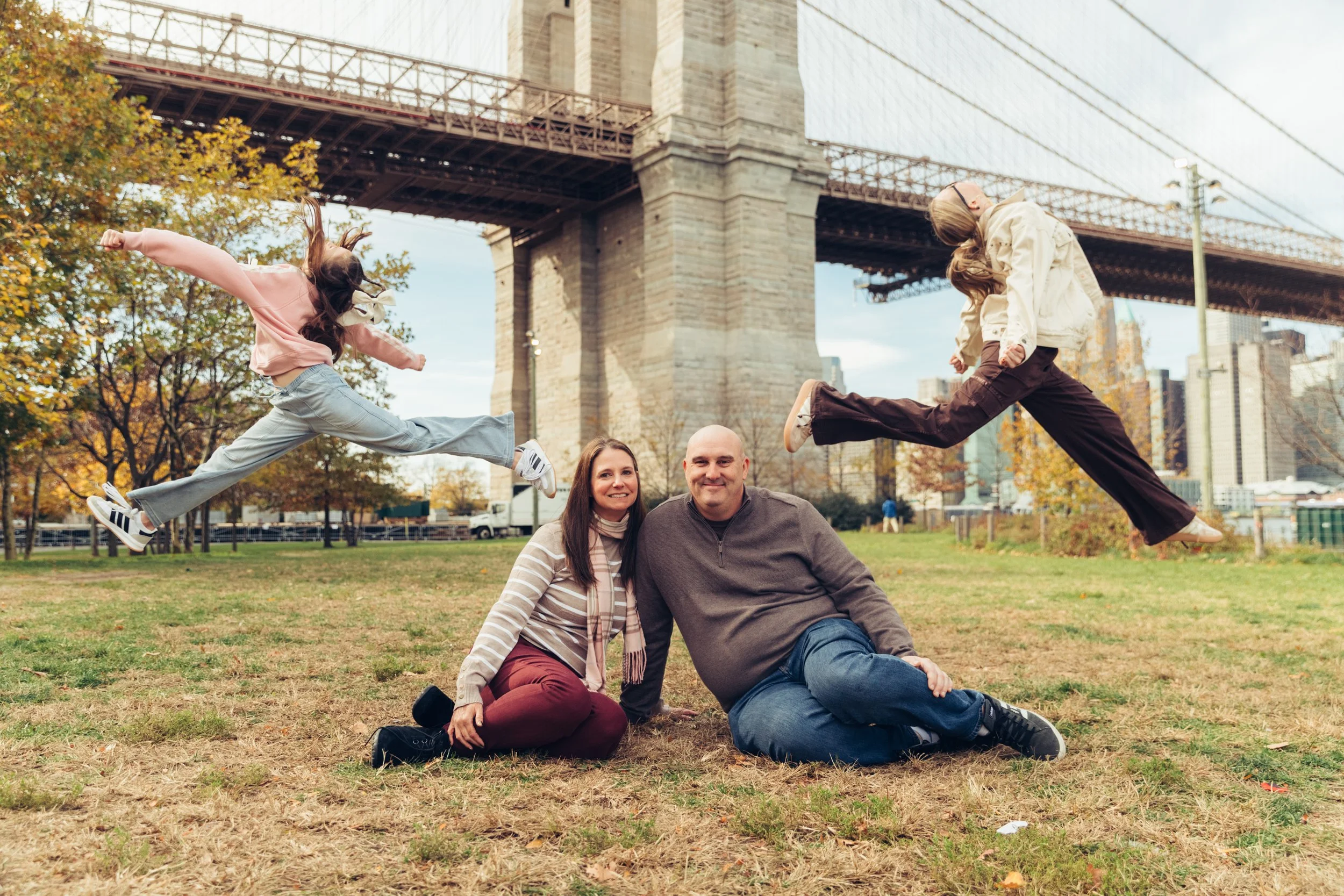 A family of four sitting and jumping in a park under the Brooklyn Bridge, with autumn trees in the background