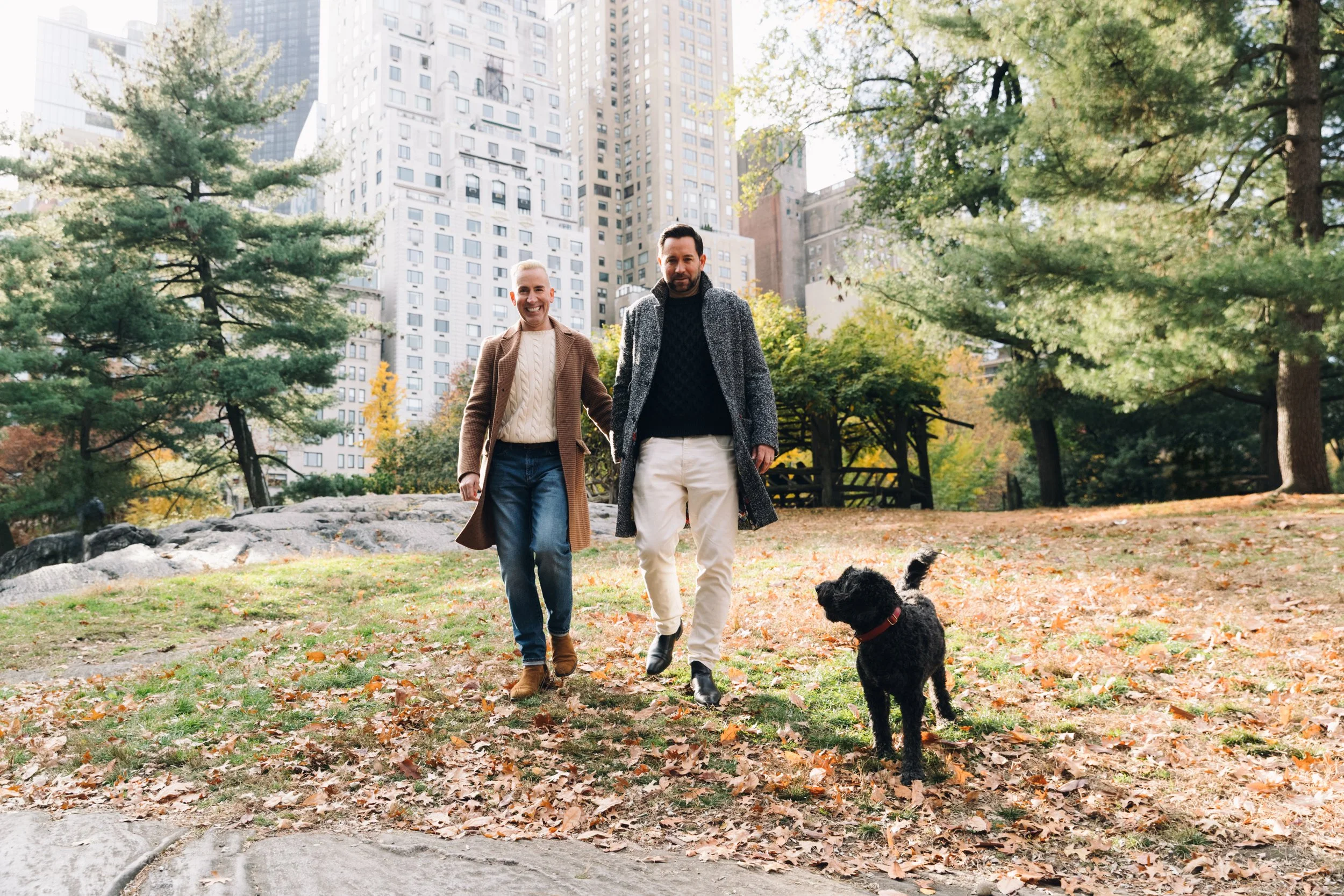Two men walking and smiling in a park with fallen leaves, greenery, trees, and tall buildings in the background, one with a black dog on a leash.