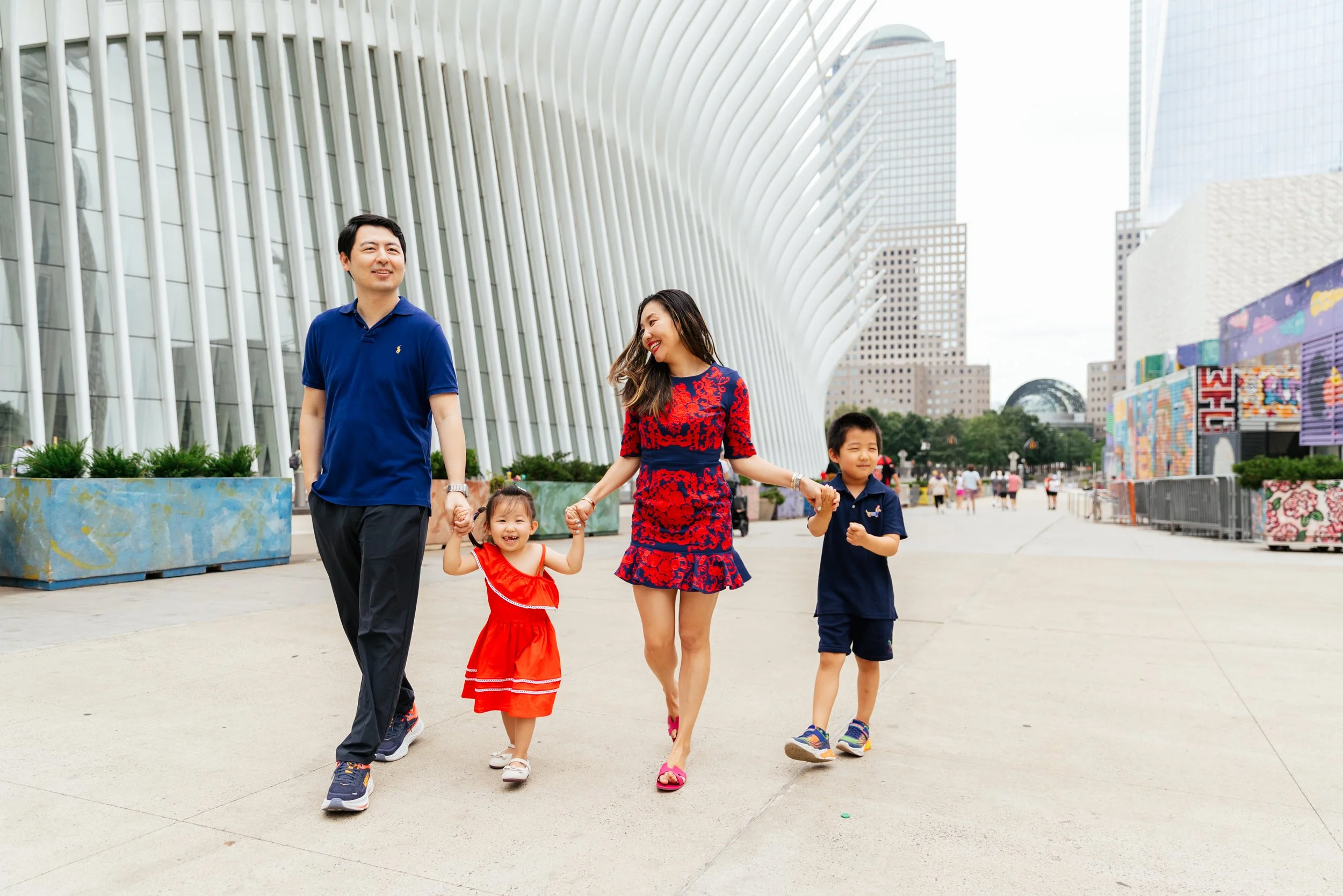 Family of four walking outdoors in an urban area with modern architecture, holding hands and smiling.