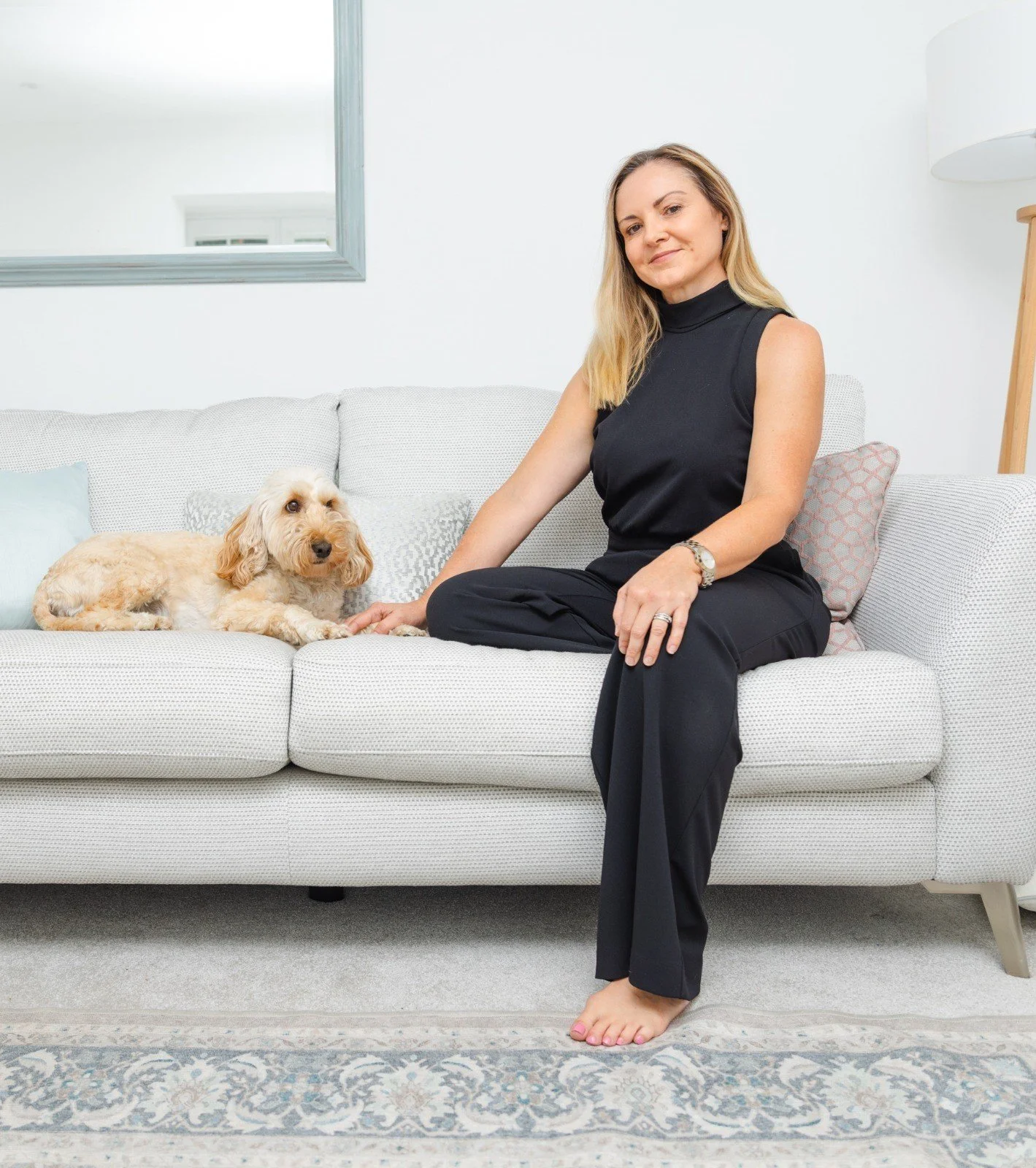 A woman sitting on a light-colored couch with a tan dog lying beside her in a bright, modern living room.