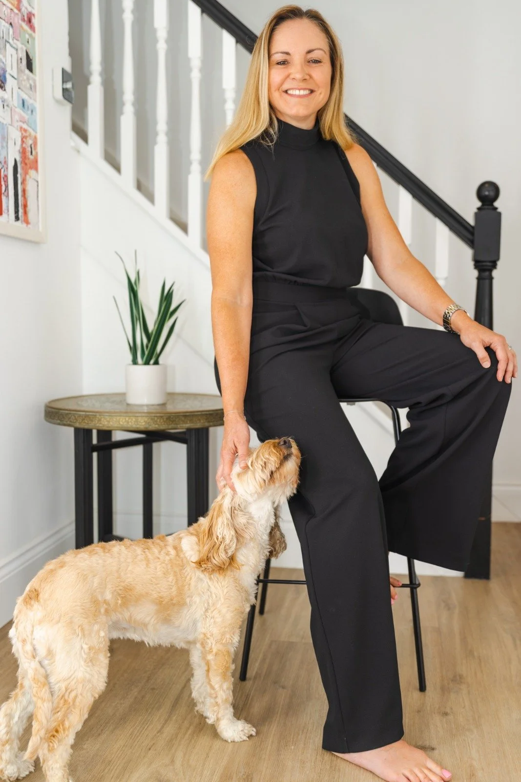 A woman in black pants and a sleeveless black top sitting on a stool, petting a curly-coated dog.