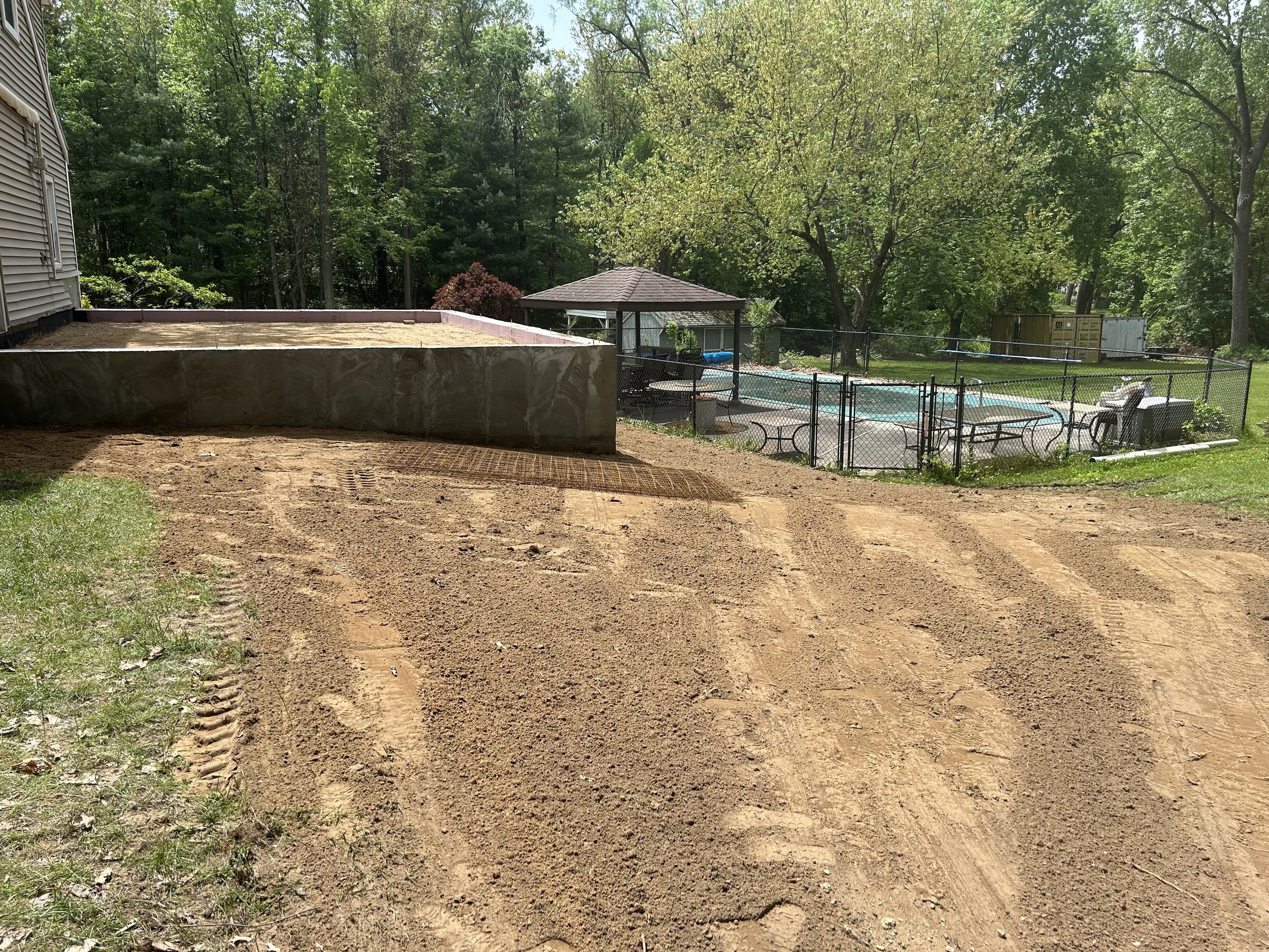 A backyard with a dirt area, a raised sandpit, a fenced pool with a gazebo, a lawn, trees, and outdoor furniture on a sunny day.