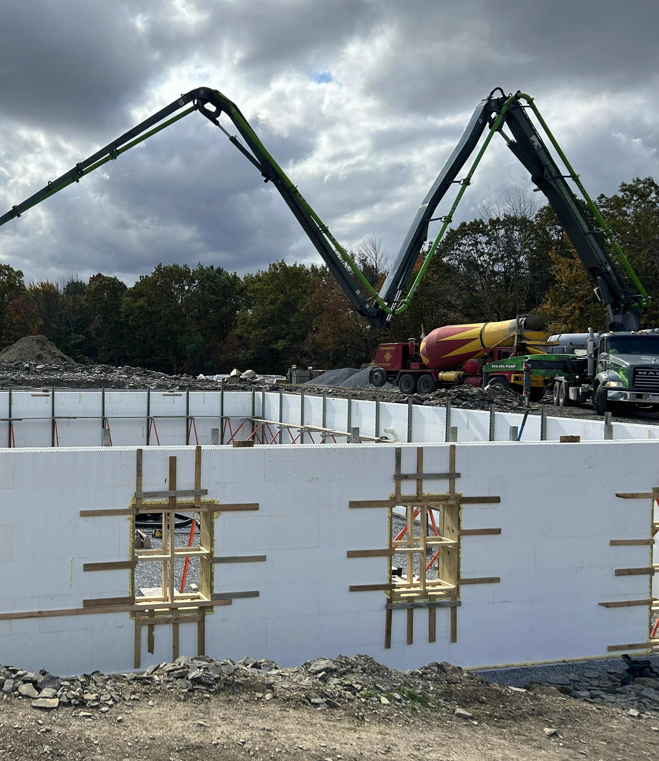 Construction site with concrete walls, window openings, and a large concrete pump truck with an extended arm, under a cloudy sky.