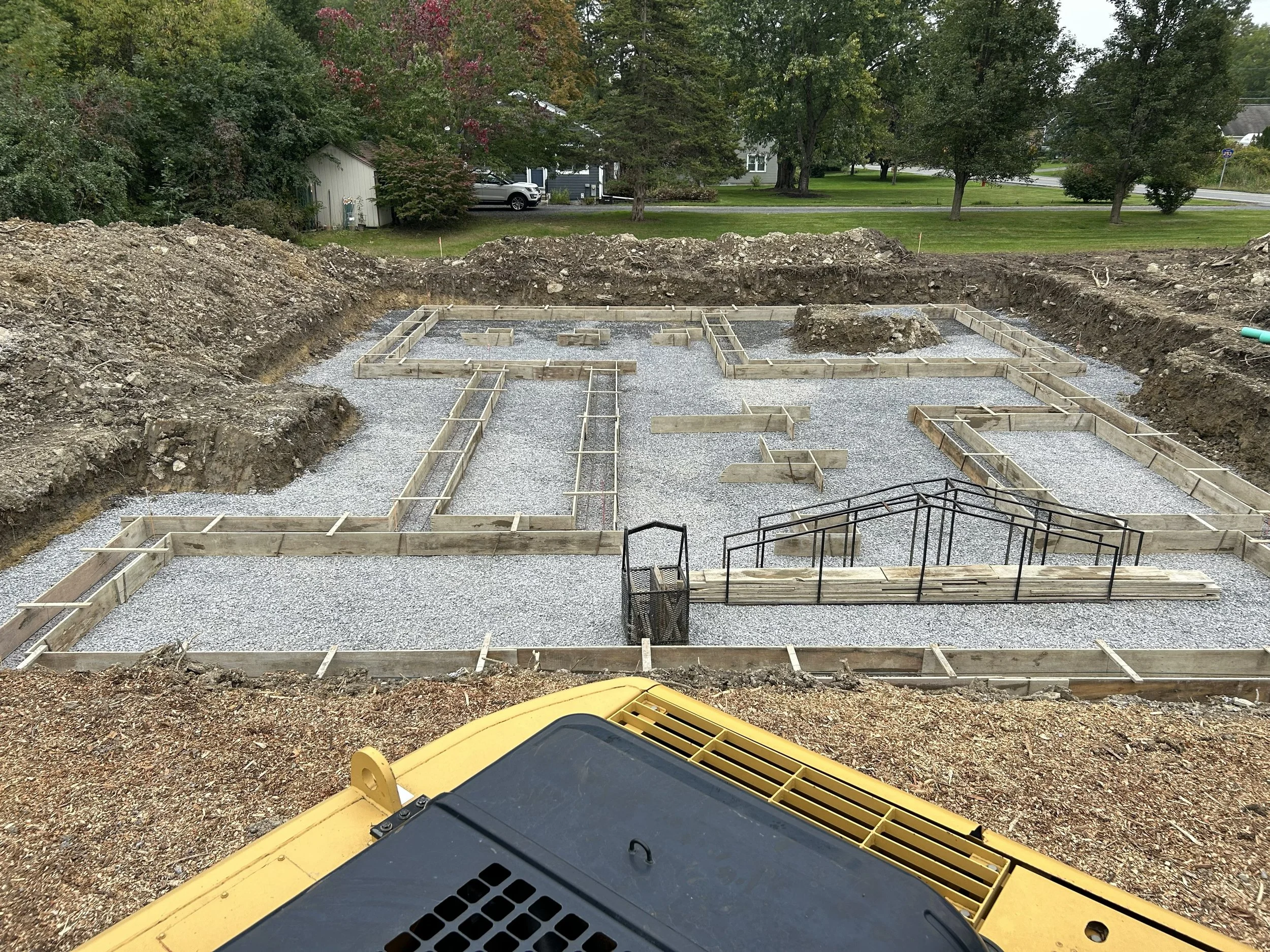 Construction site with foundation framing for a building, gravel base, and some wooden and metal supports, with trees and houses in the background.