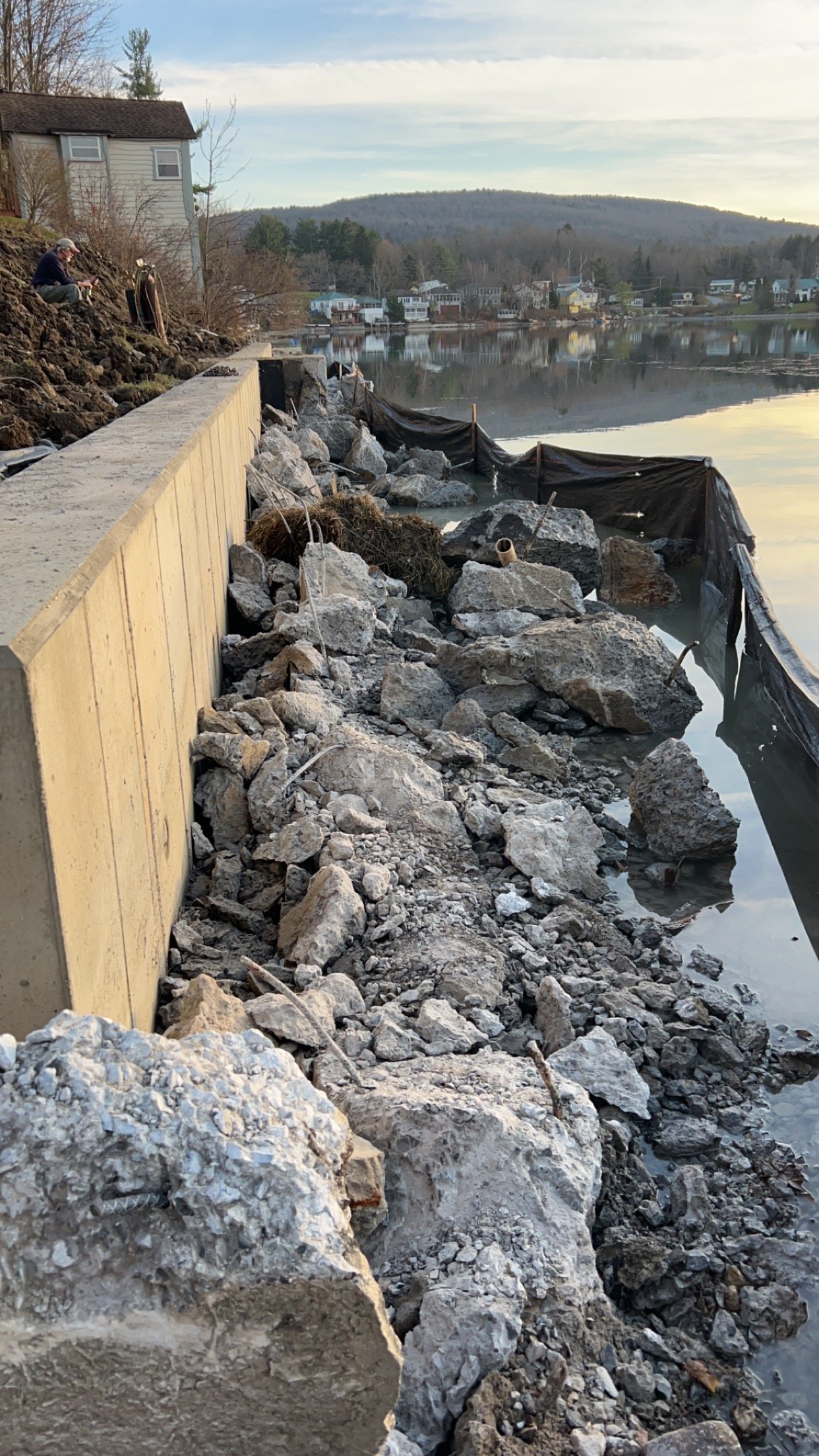 A damaged lakeside shoreline with large rocks and a bent black drainage pipe, a wooden retaining wall, and a calm lake with houses in the distance.