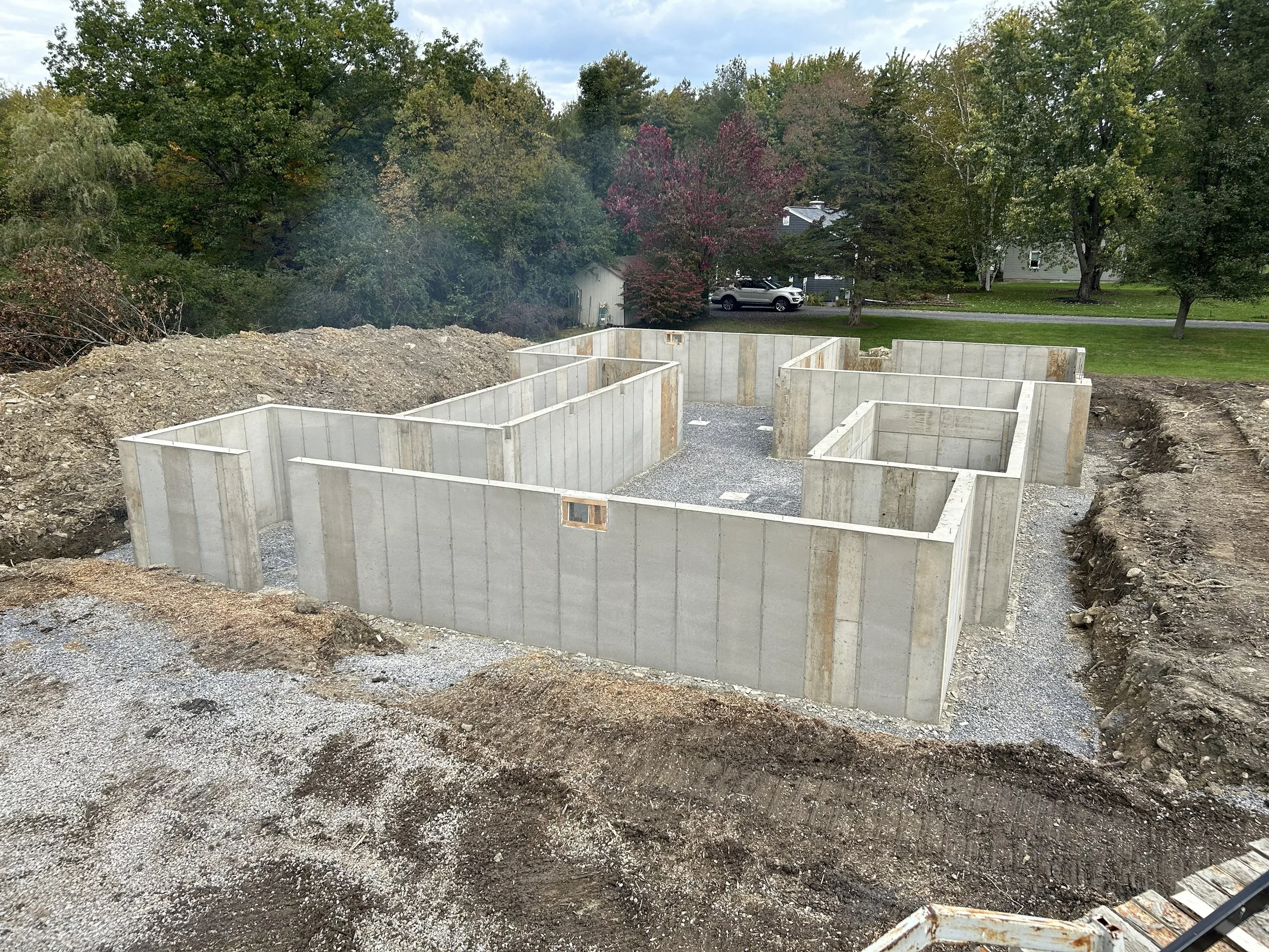 Foundation for a building under construction with concrete walls on a gravel base, surrounded by dirt and soil, with trees and houses in the background.