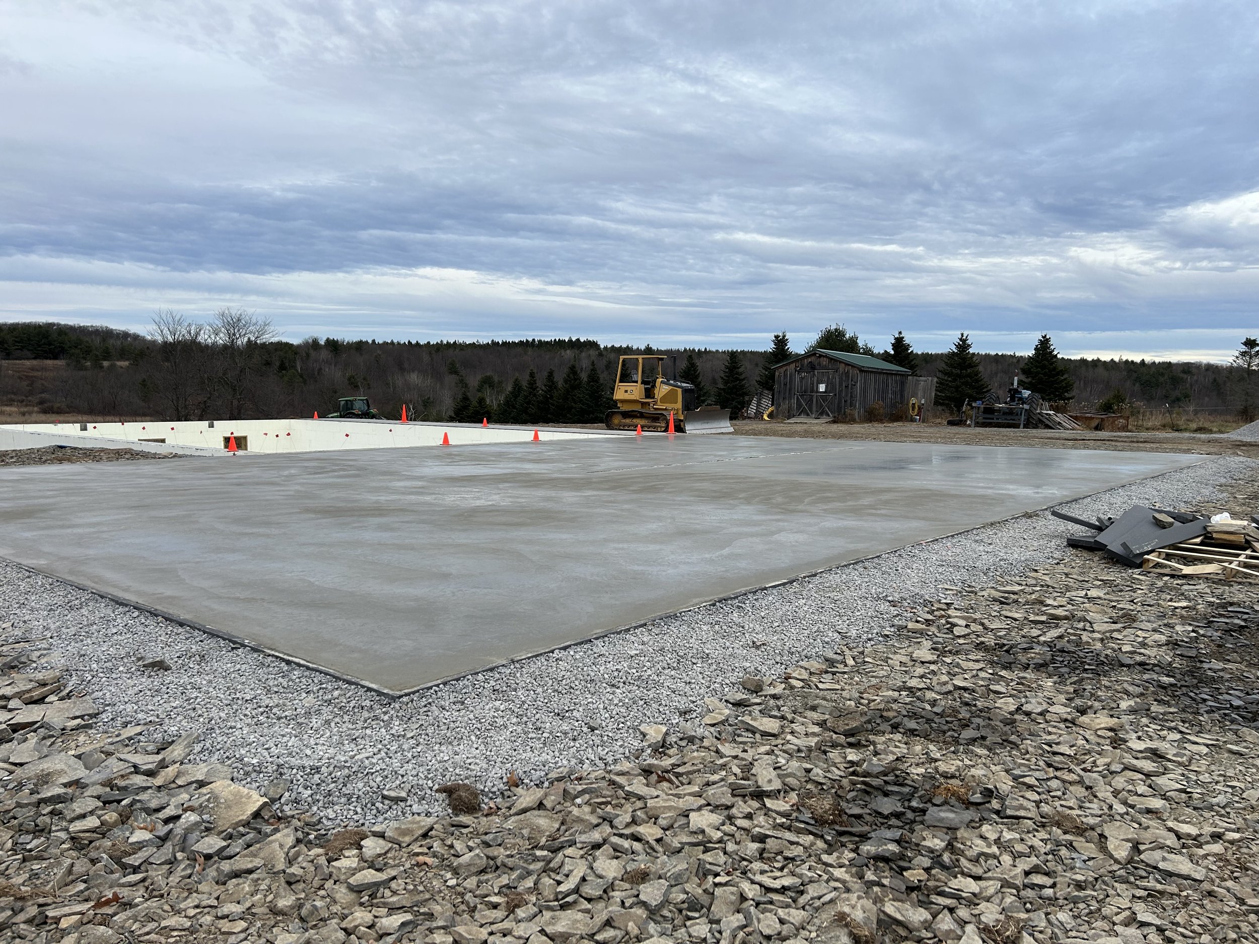 Construction site with a lowered concrete foundation, construction equipment, and a wooden shed in the background under a cloudy sky.
