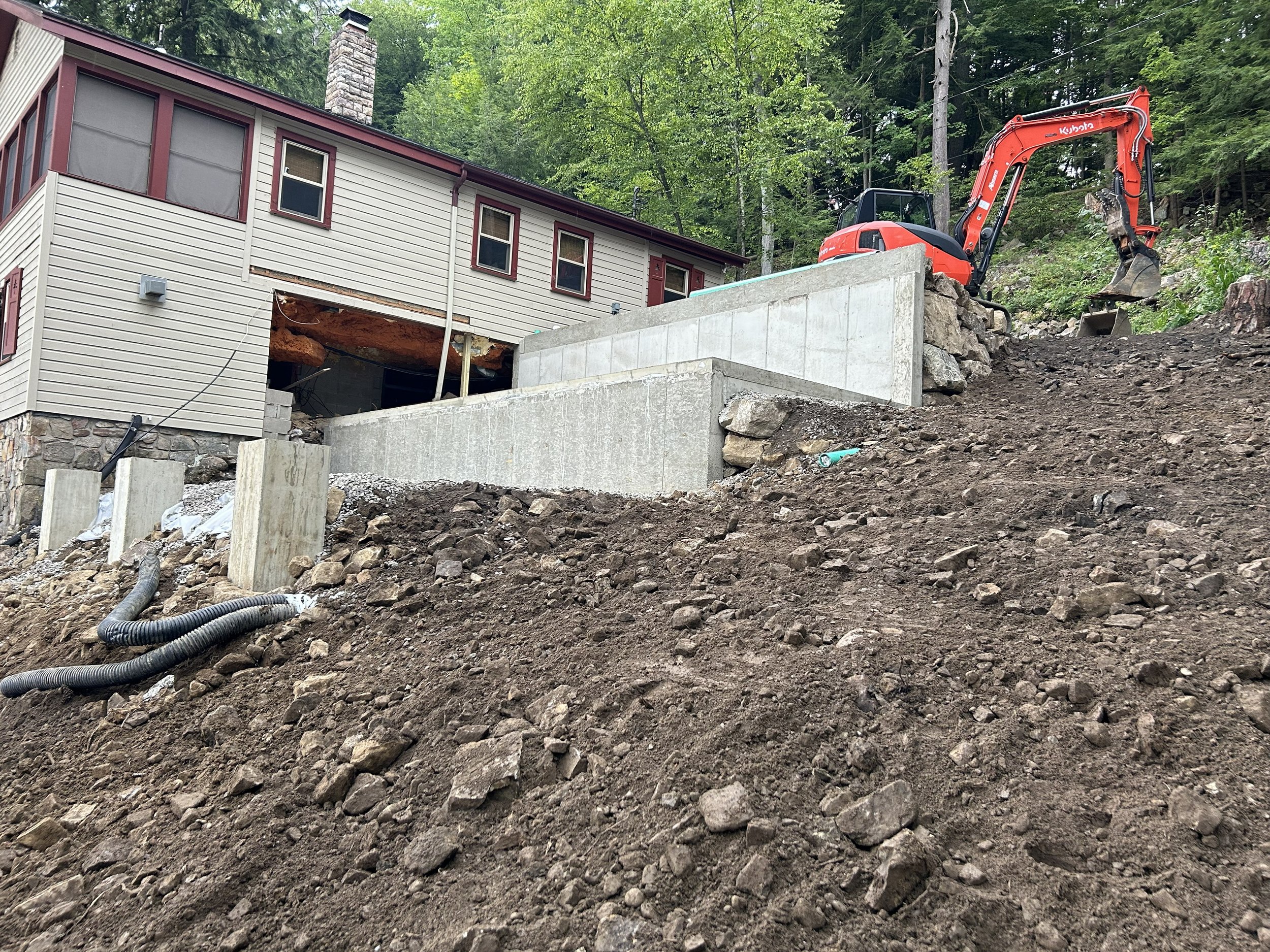Construction site on a hillside with a small excavator on the right, working near a house that is partially built into the slope. Visible dirt and rocks in the foreground, concrete retaining walls, and construction materials scattered around.