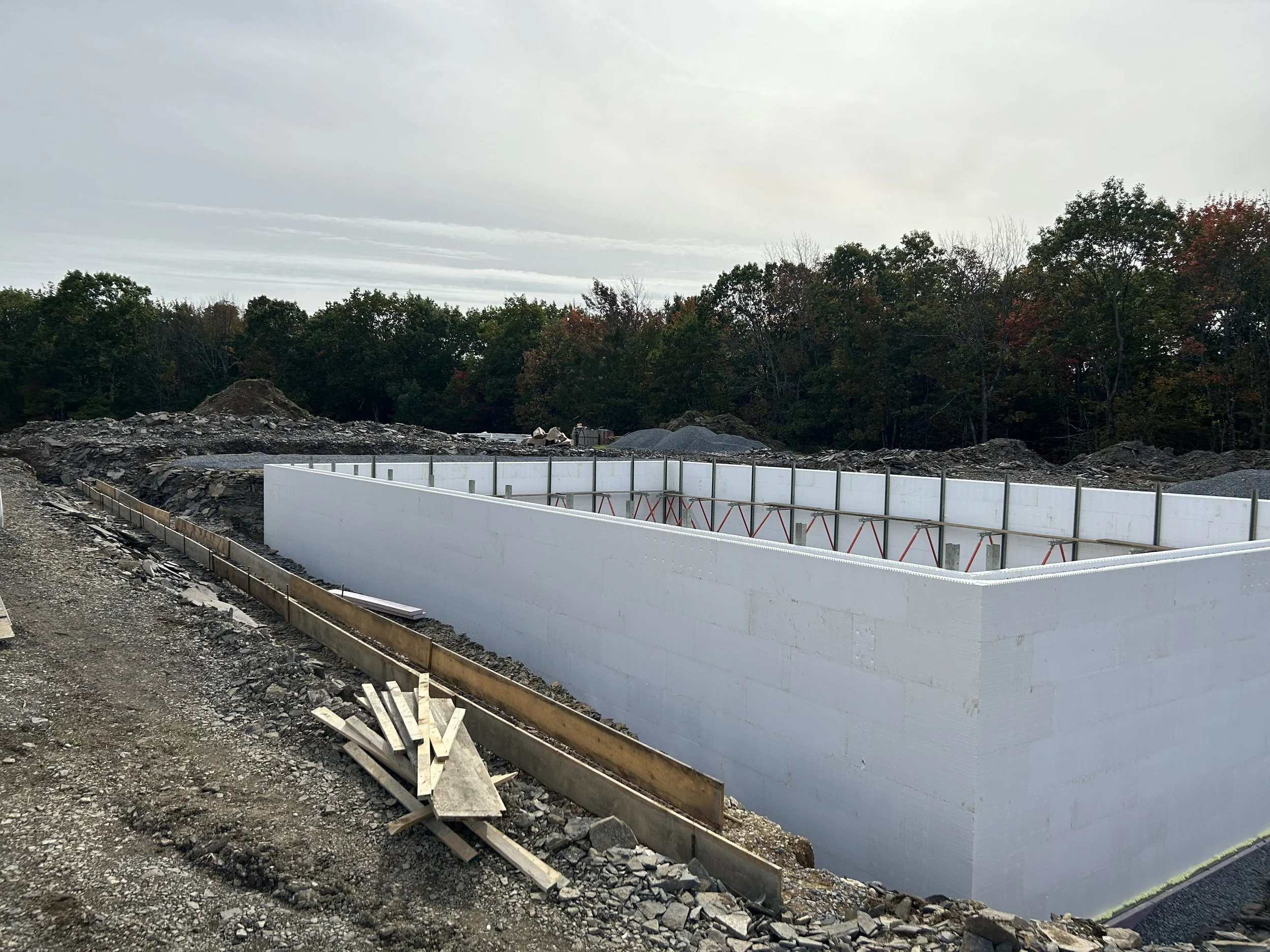 Construction site with a white foundation wall and piles of gravel, surrounded by trees and an overcast sky.