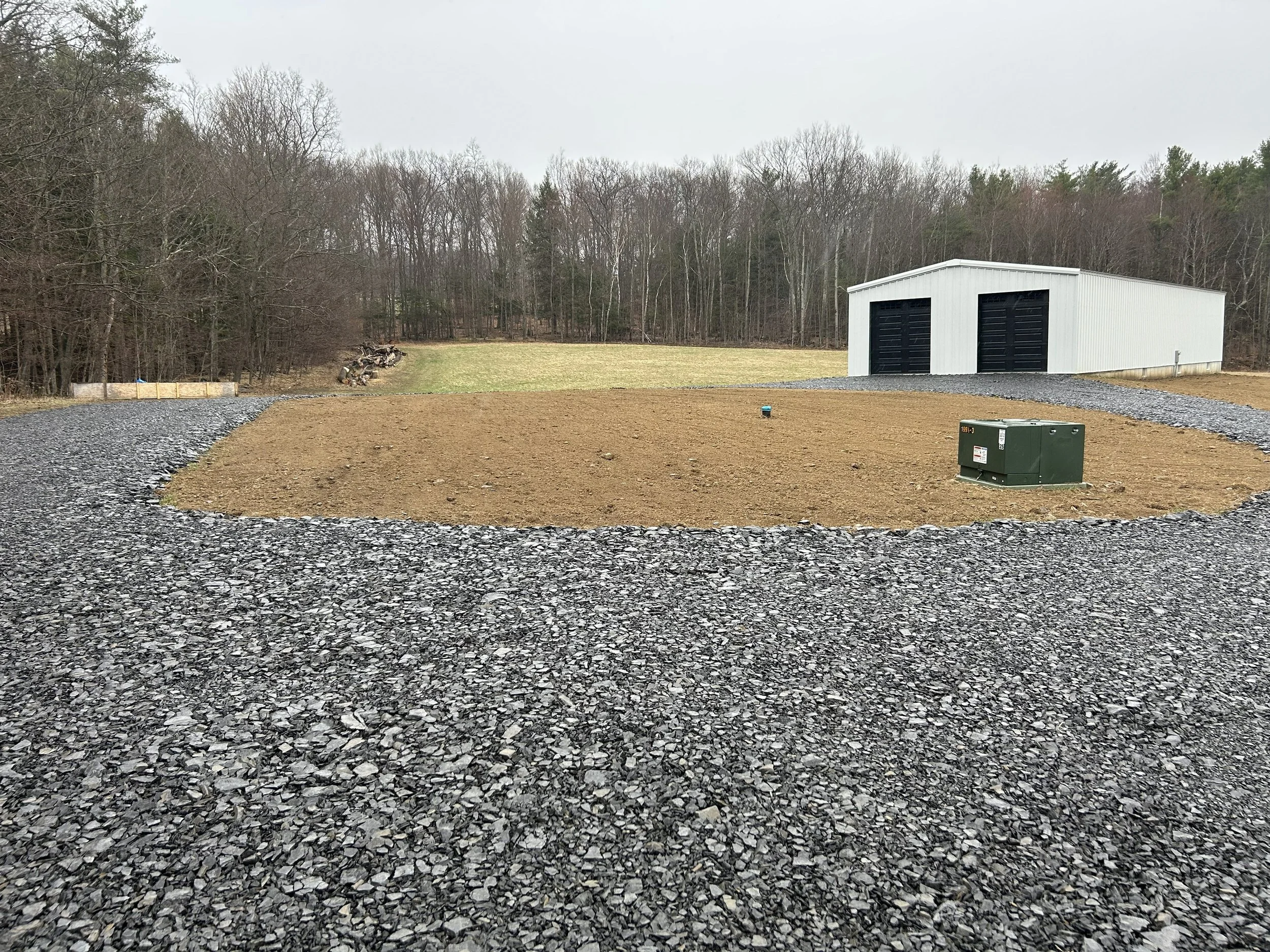 A gravel driveway surrounds a bare patch of dirt in front of a white storage building with black doors, located in a rural area with trees in the background.