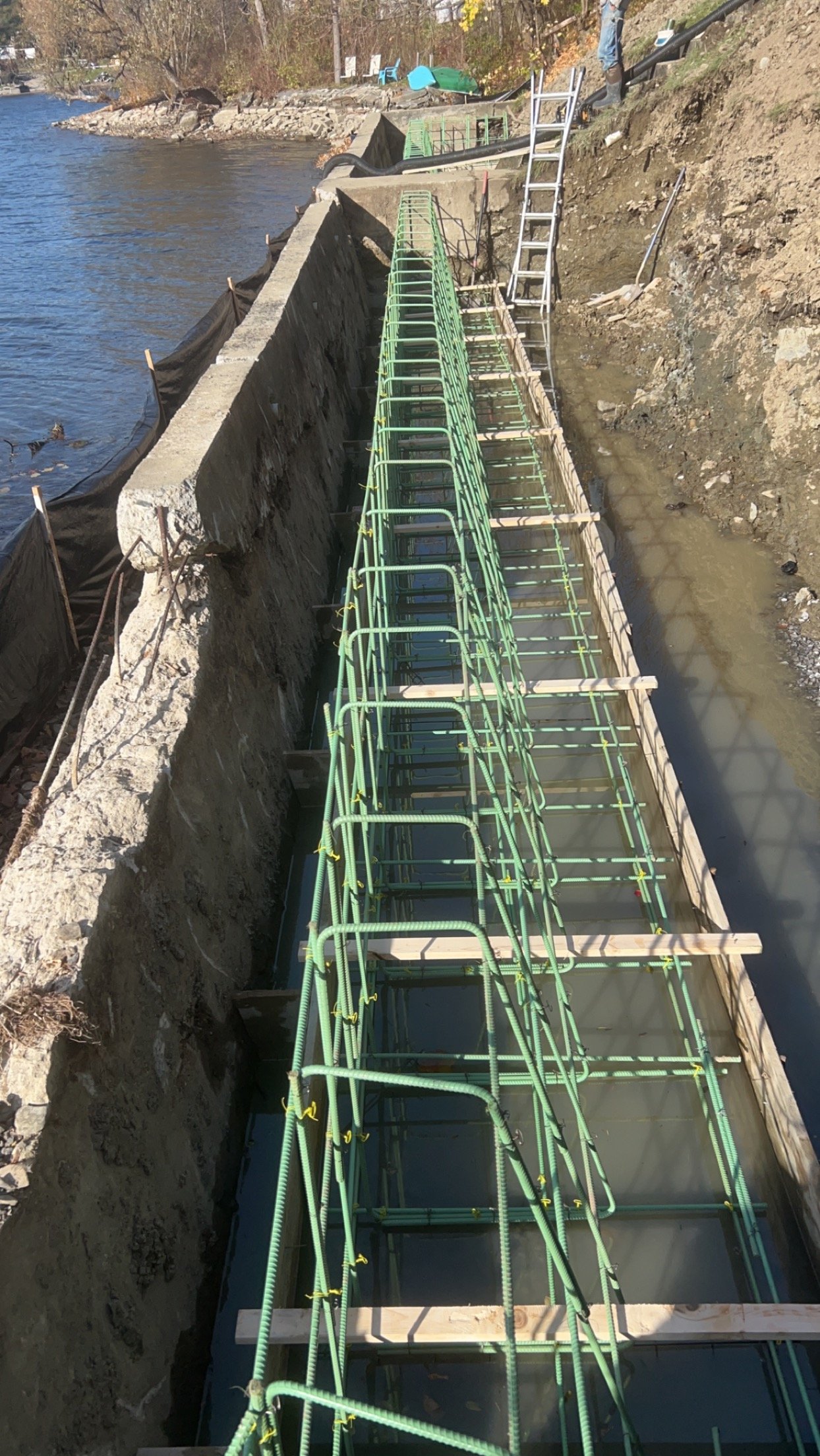 Under construction bridge with green rebar and wooden formwork along a riverbank.