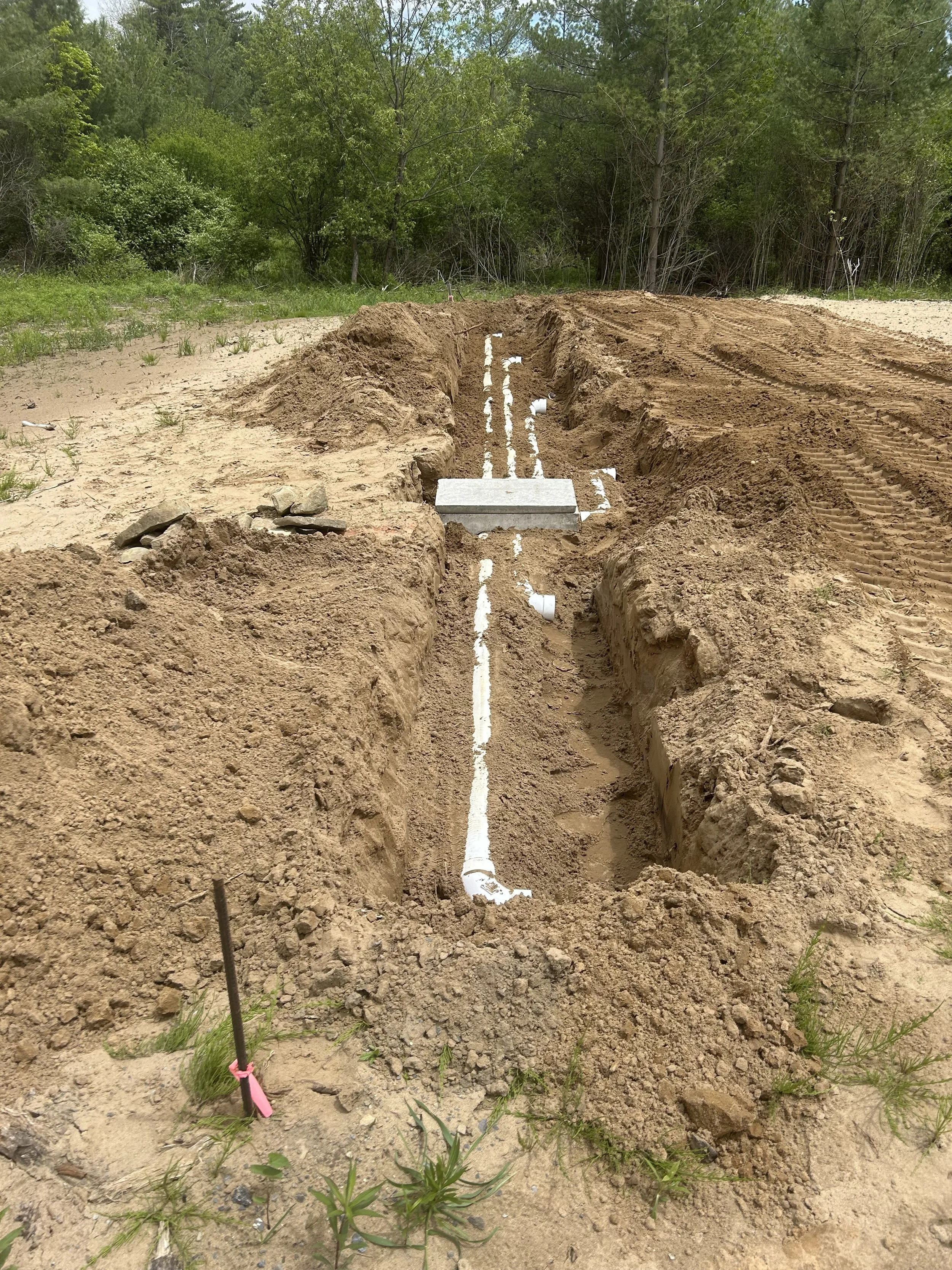 Underground irrigation pipes being installed in a dirt trench at a construction site surrounded by trees.