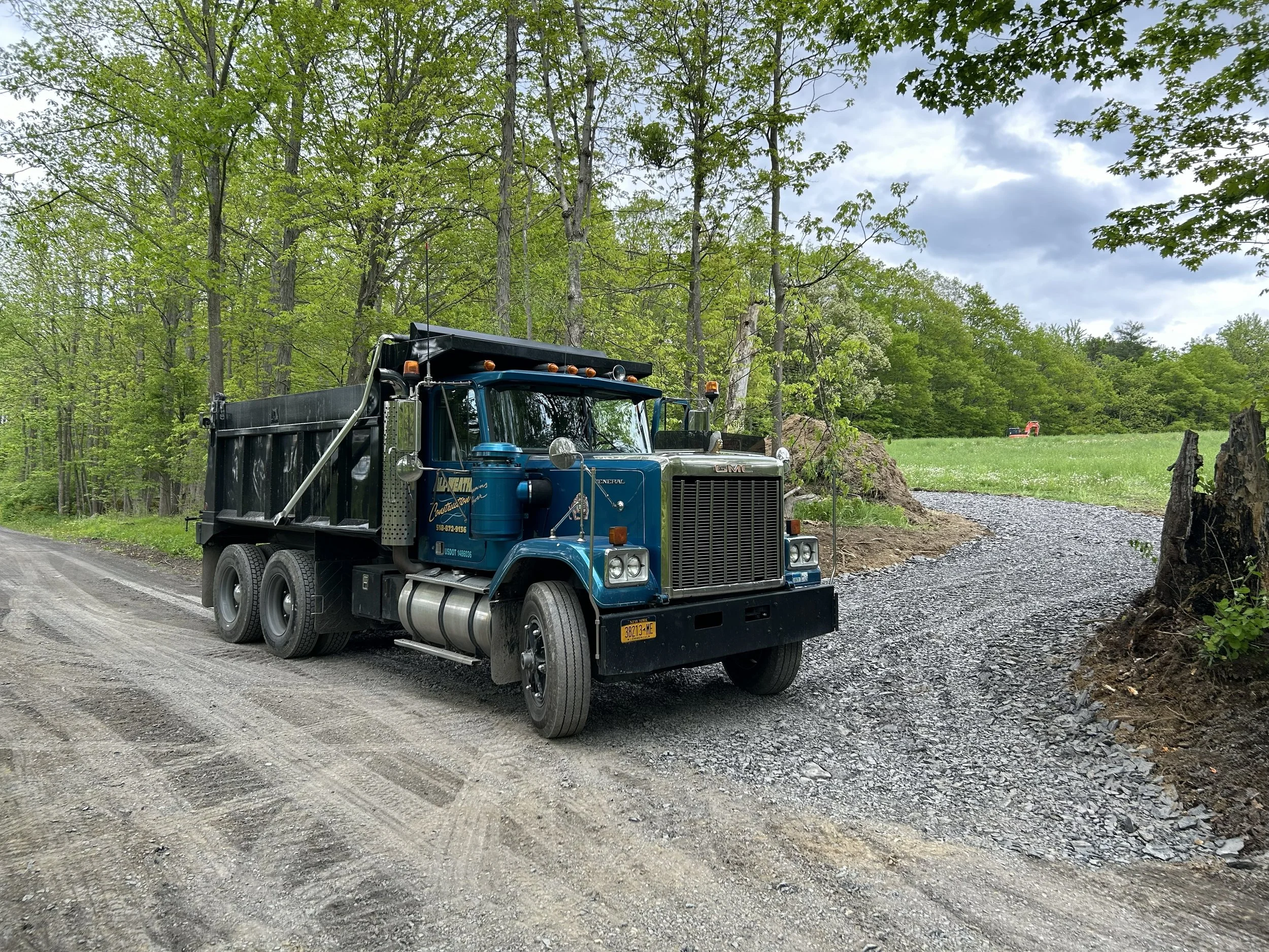 A blue GMC dump truck parked on a gravel road surrounded by green trees under a cloudy sky.
