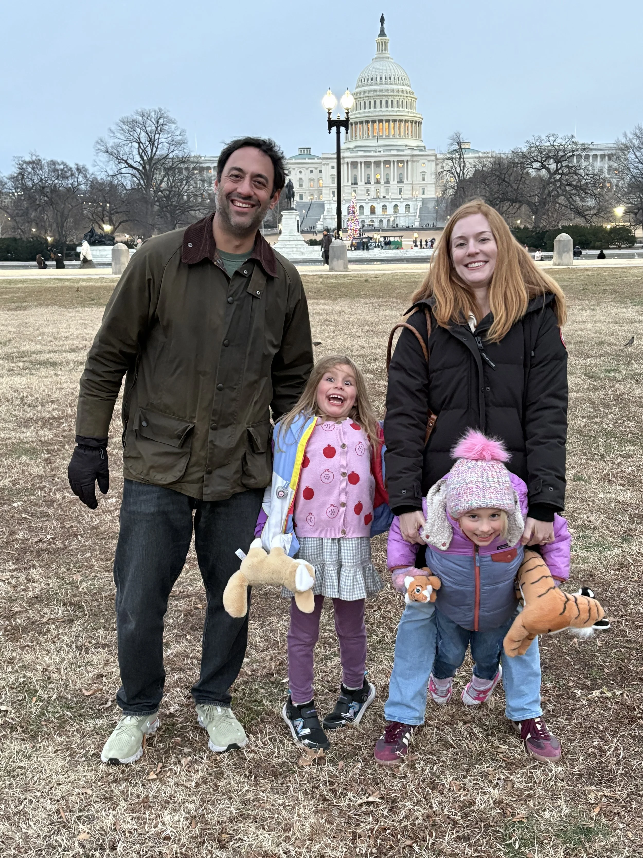 A family of four standing on a field in front of the US Capitol building during winter. The two children are holding plush animal toys, and all are smiling.
