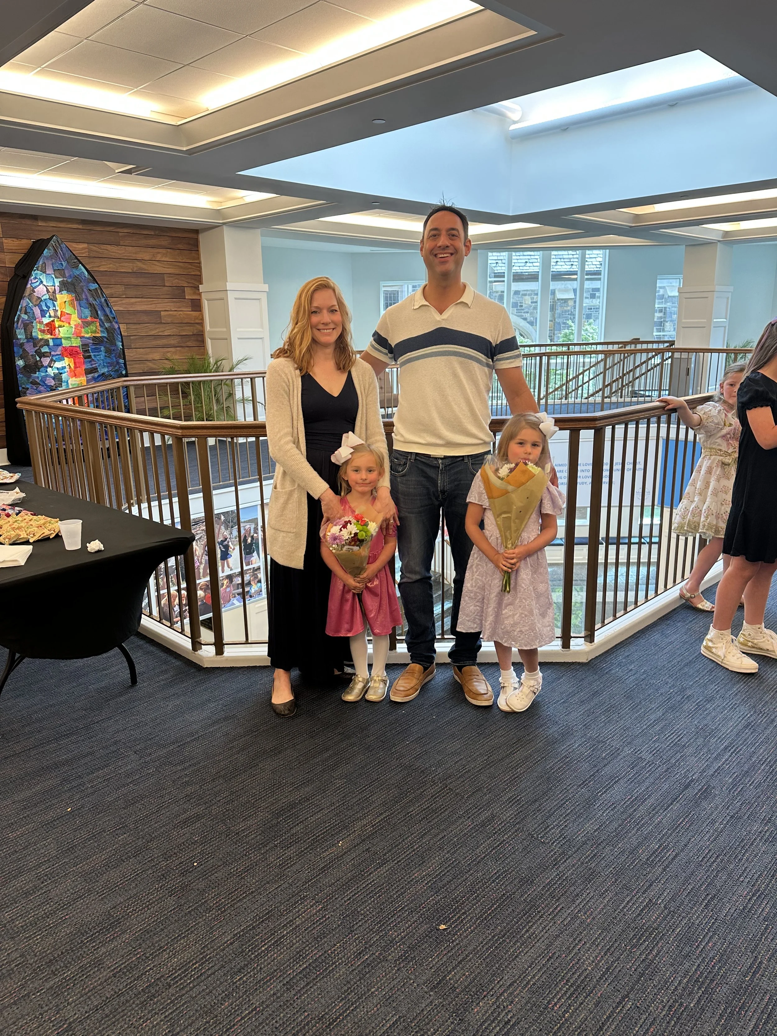 A family of four standing together at a social gathering inside a building with a view of a balcony and windows in the background. The family includes two young girls holding bouquets, a woman, and a man.